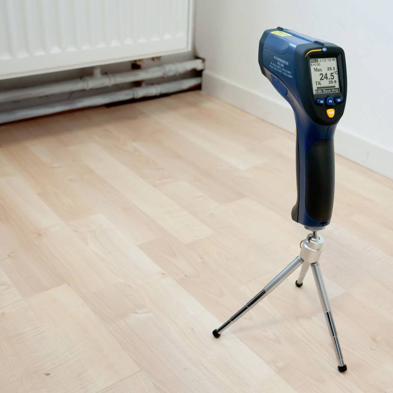 Ein Infrarot-Temperaturmessgerät mit Stativ steht auf einem Holzboden neben einem Heizkörper. Das Display zeigt 24,5°C an.