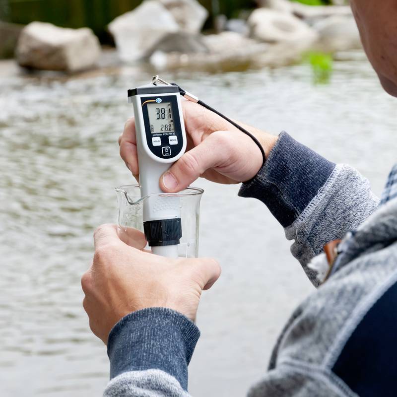 Eine Person misst die Wassertemperatur eines Teichs mit einem digitalen Thermometer, das 20,8 Grad Celsius anzeigt.
