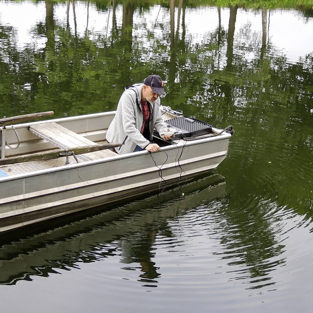 Ein Mann sitzt in einem kleinen Boot auf einem ruhigen See und arbeitet mit elektronischen Geräten, umgeben von Bäumen am Ufer.