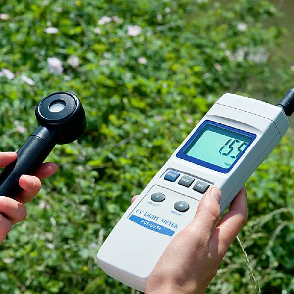 Zwei Hände halten ein digitales Messgerät im Grünen, das einen UV-Wert von 0,55 anzeigt. Ein Sensor ist mit einem Kabel verbunden.