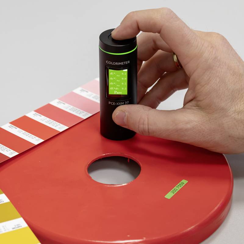 Eine Hand hält ein Kolorimeter, das auf einer Farbmusterkarte steht. Das Kolorimeter zeigt Werte an, die zur Farbmessung verwendet werden.