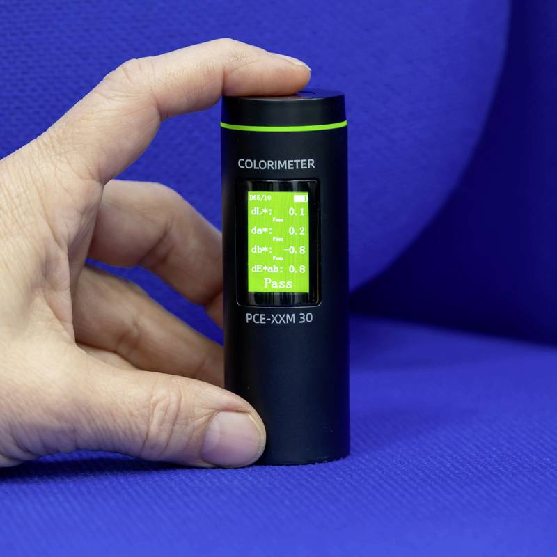 Eine Hand hält ein tragbares Kolorimeter mit grün leuchtendem Display, das Messwerte anzeigt. Das Gerät dient zur Farbanalyse.