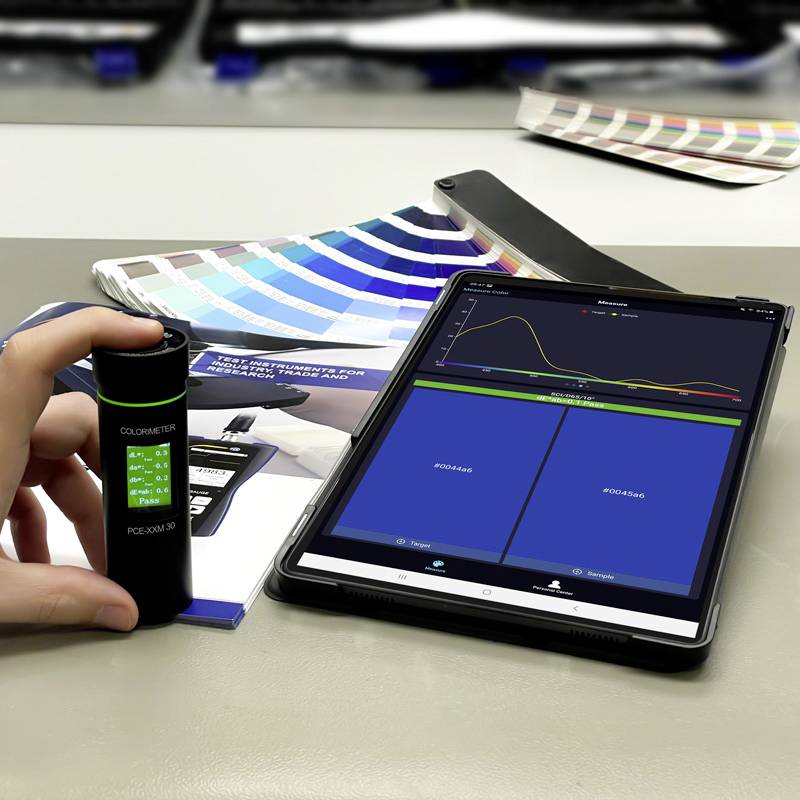 Hand hält ein Colorimeter neben einem Tablet. Auf dem Tablet wird ein Farbmessdiagramm angezeigt. Farbmusterfächer im Hintergrund.