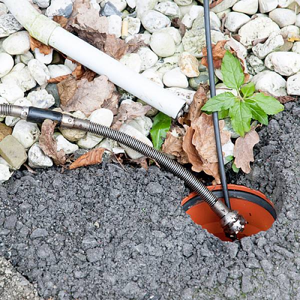 Abflussreinigungswerkzeug in einem Loch mit Asphalt und Kies. Blätter und eine Pflanze sind zu sehen.