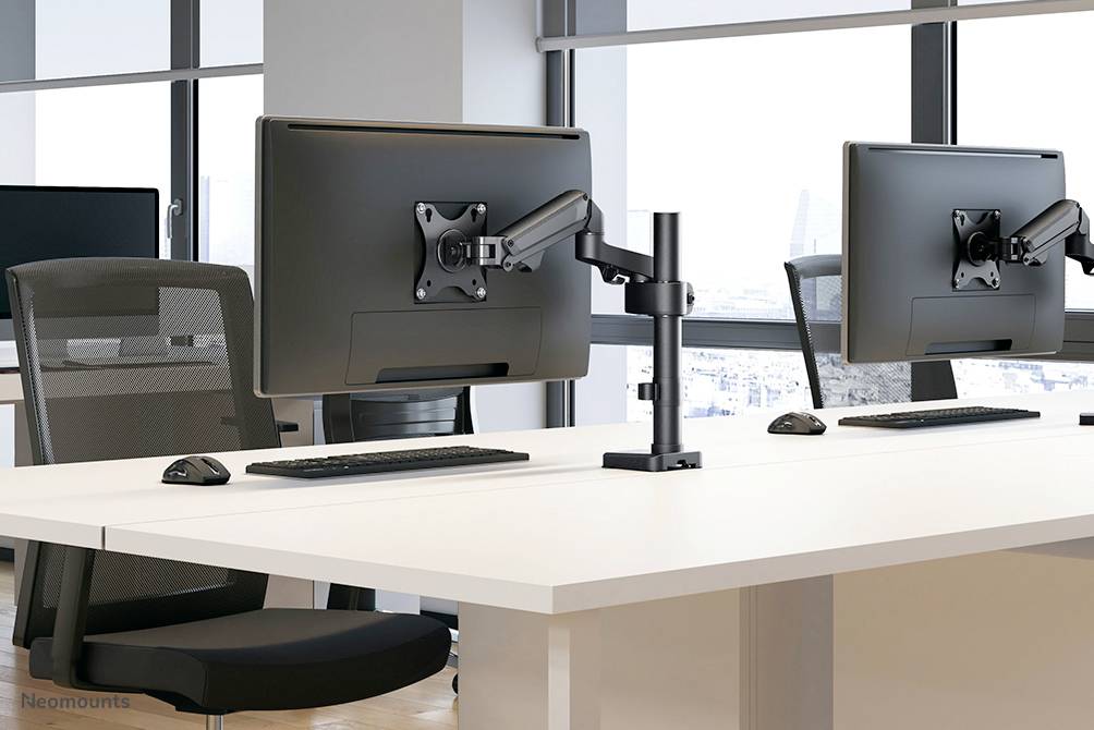 Zwei moderne Monitore auf weißen Schreibtischen in einem Büro mit großen Fenstern. Daneben sind Bürostühle zu sehen, helle und geräumige Umgebung.