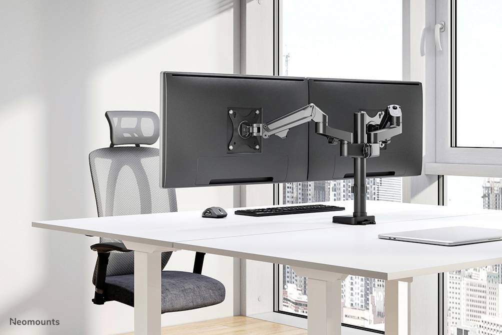 Ein moderner, minimalistischer Büroschreibtisch mit zwei Computerbildschirmen an einem dualen Monitorarm. Ein ergonomischer Stuhl steht davor. Im Hintergrund sind große Fenster mit Stadtblick.