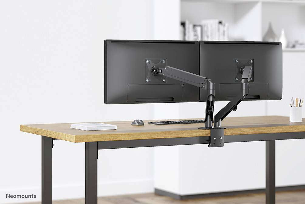 Ein moderner Schreibtisch mit zwei nebeneinander montierten Monitoren auf einem verstellbaren Arm, eine Tastatur darunter und eine Tasse rechts.