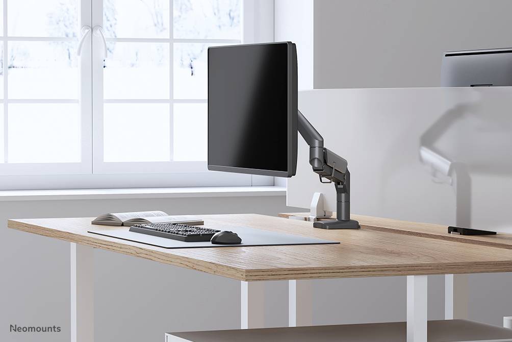Ein modernes Büro mit einem Schreibtisch, Monitor auf Halterung, Tastatur und Maus. Große Fenster im Hintergrund lassen Licht herein.
