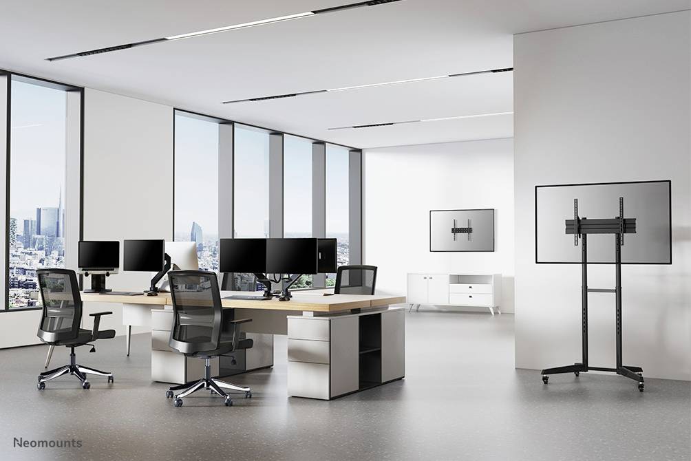 Ein modernes Büro mit einem großen Schreibtisch, mehreren Monitoren und ergonomischen Stühlen. Fenster bieten Blick auf eine Stadtlandschaft.