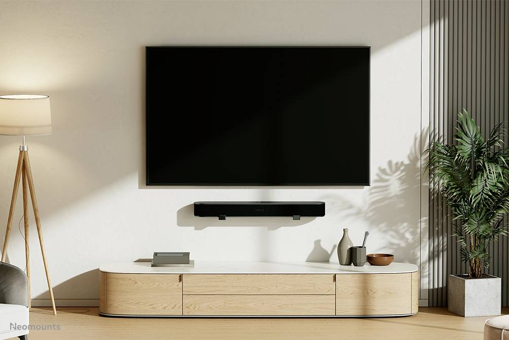 Moderne Wohnzimmer-Einrichtung mit Wandfernseher, darunter eine Soundbar und ein Holzschrank. Eine Lampe links und eine Pflanze rechts.