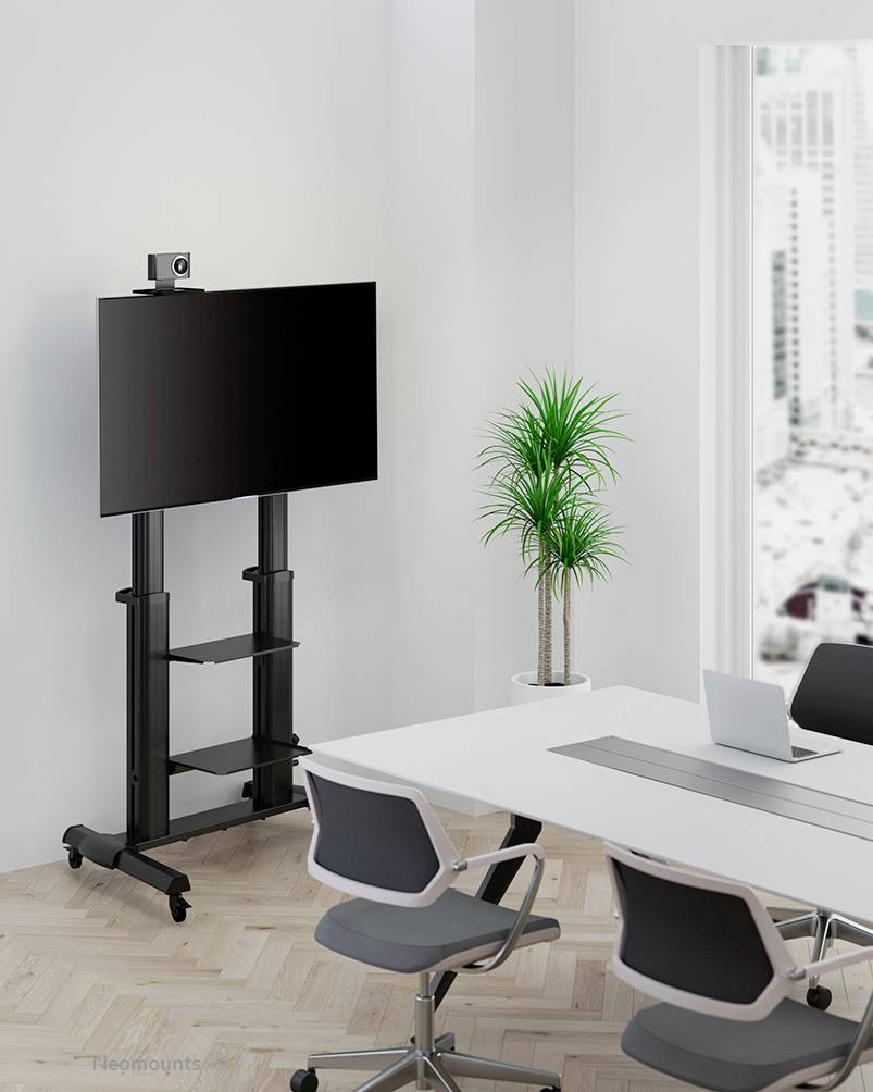 Ein moderner Konferenzraum mit weißem Tisch, drei Bürostühlen und einem großen Flachbildschirm auf einem fahrbaren Ständer. Im Hintergrund steht eine grüne Pflanze.