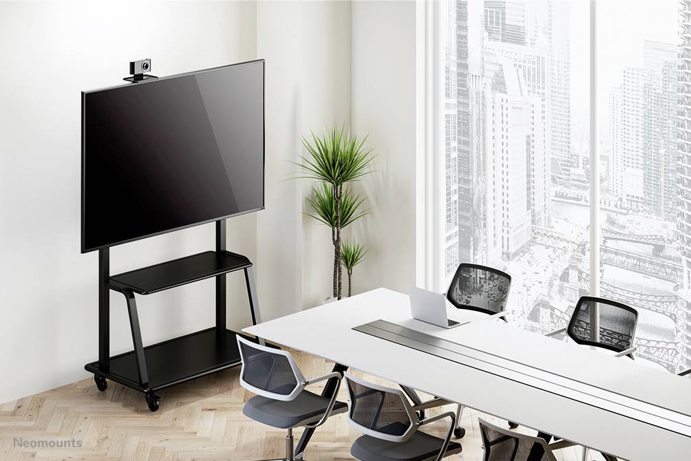 Der Konferenzraum zeigt einen großen Bildschirm auf einem Rollwagen, einen langen Tisch mit schwarzen Stühlen und ein Fenster mit Stadtblick.