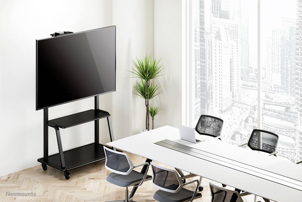 Ein moderner Konferenzraum mit einem großen Bildschirm auf einem mobilen Ständer, einem weißen Tisch und vier Stühlen vor einer Fensterfront.