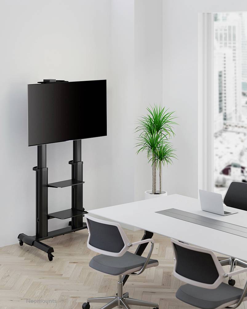 Ein moderner Konferenzraum mit großem Monitor auf einem Ständer, weißen Tisch und drei Bürostühlen. Im Hintergrund steht eine Topfpflanze.