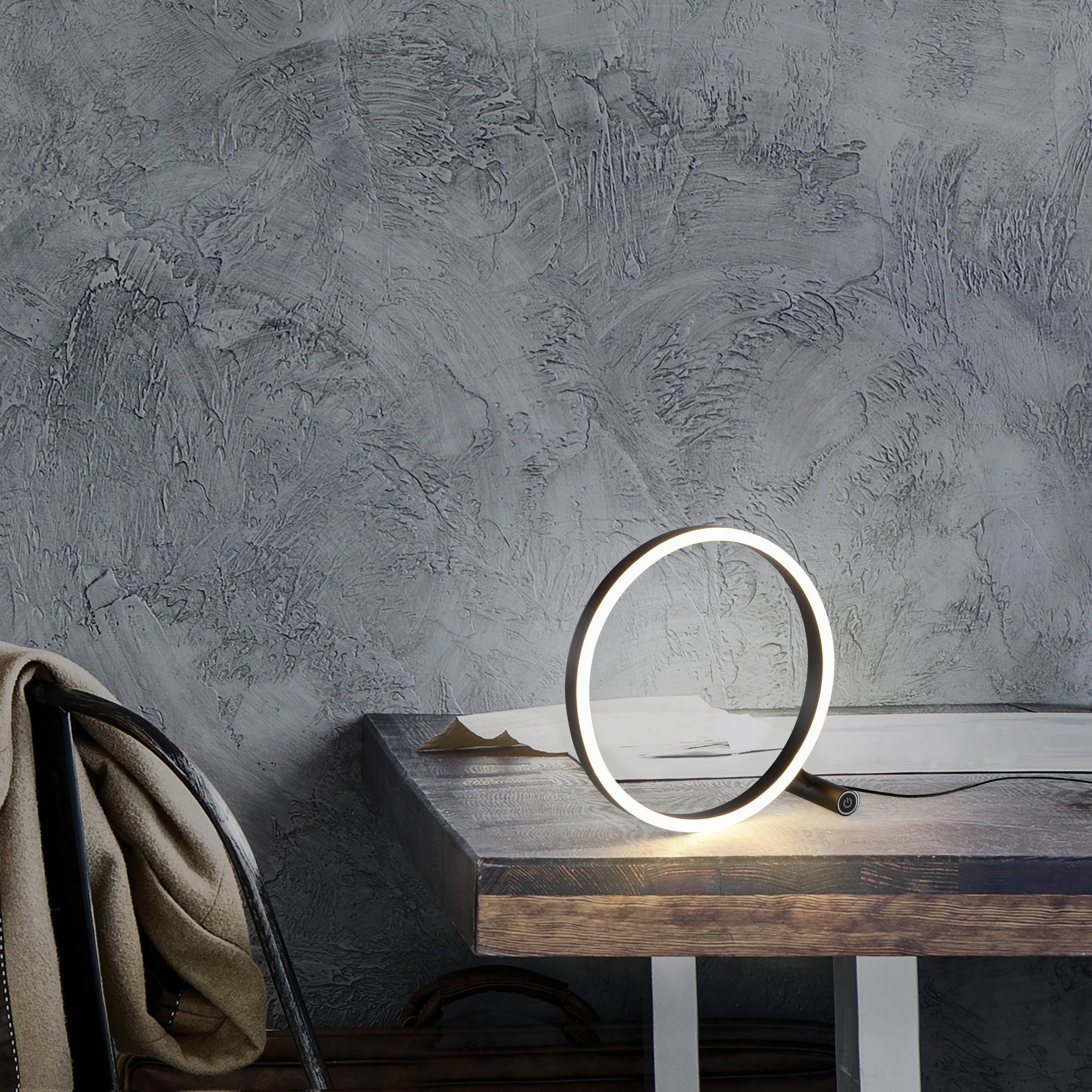 Eine runde, leuchtende Tischlampe steht auf einem Holztisch vor einer grauen, strukturierten Wand. Daneben liegt ein beiger Schal auf einem Stuhl.
