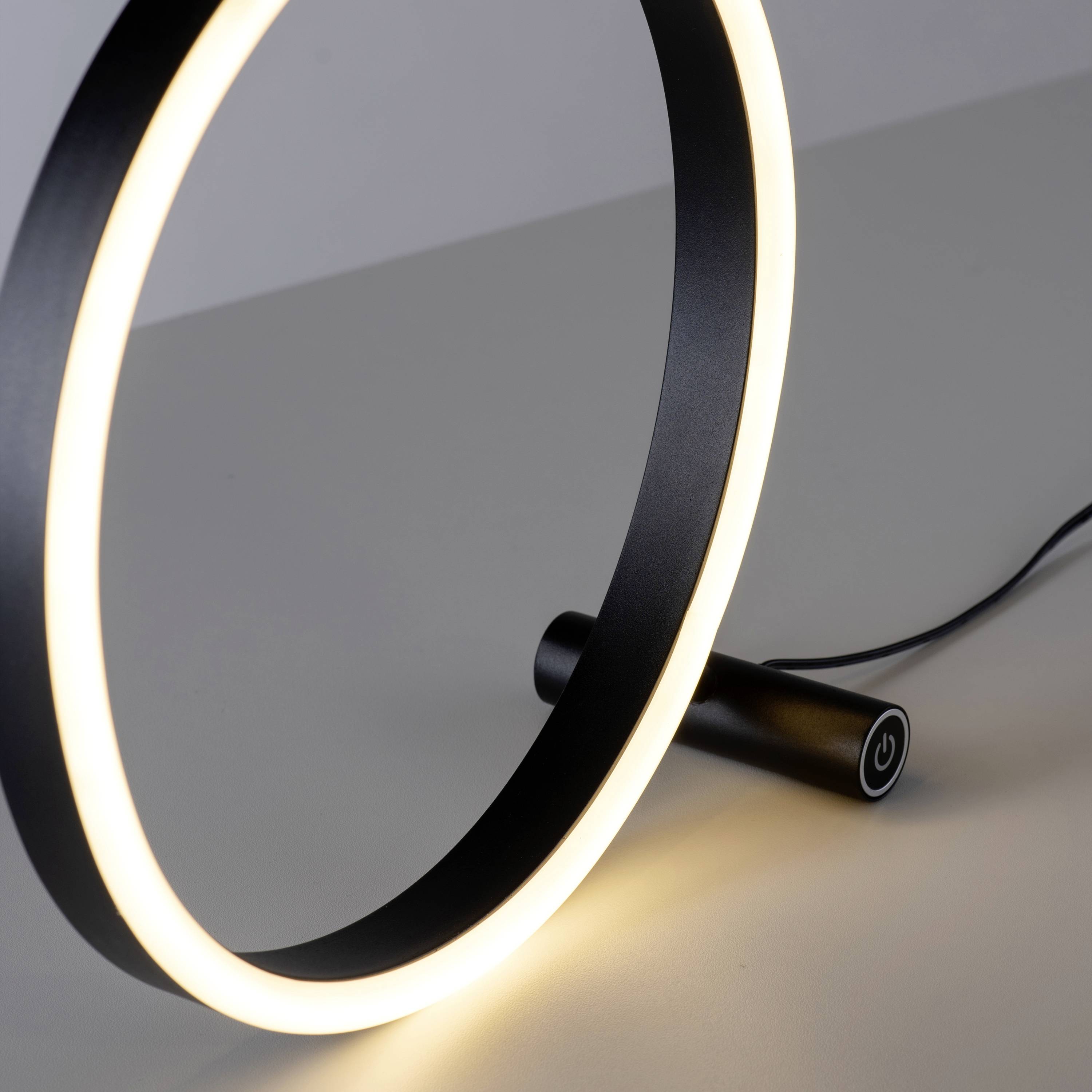 Eine moderne Tischlampe in Ringform leuchtet auf einem weißen Tisch. Der runde Ein-/Ausschalter ist benutzerfreundlich gestaltet.