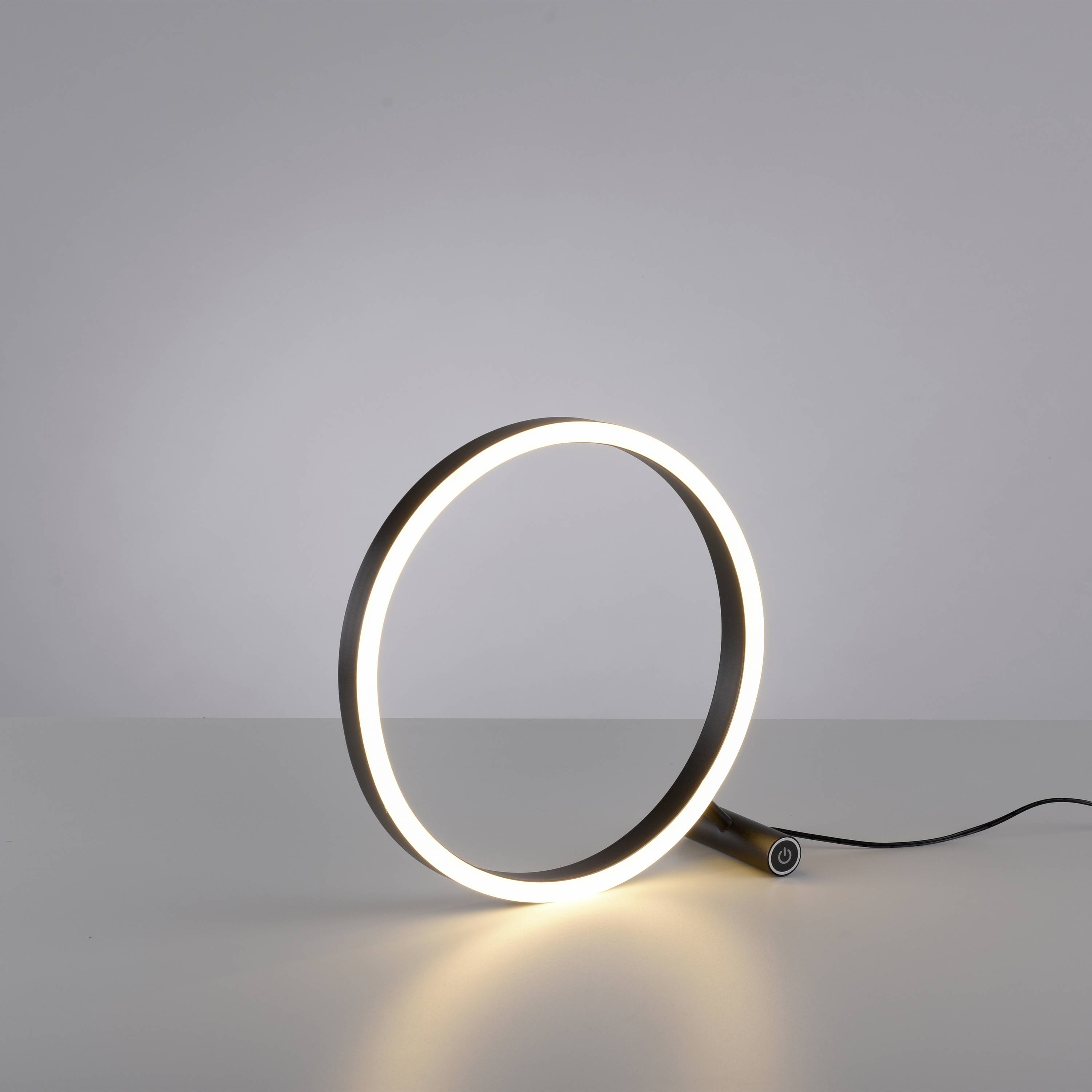 Eine moderne Tischlampe in Form eines leuchtenden Rings steht auf einem weißen Tisch vor einem neutralen Hintergrund.