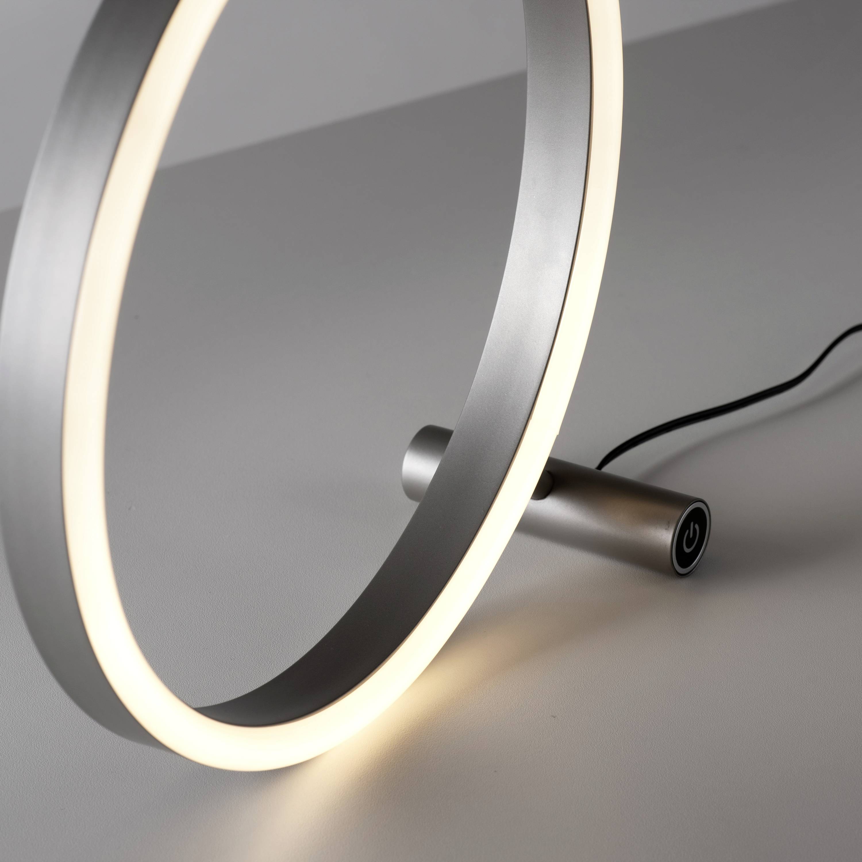 Ein moderner Lampenring aus Metall mit leuchtendem Streifen, daneben ein Schalter mit Kabel auf einem grauen Hintergrund.