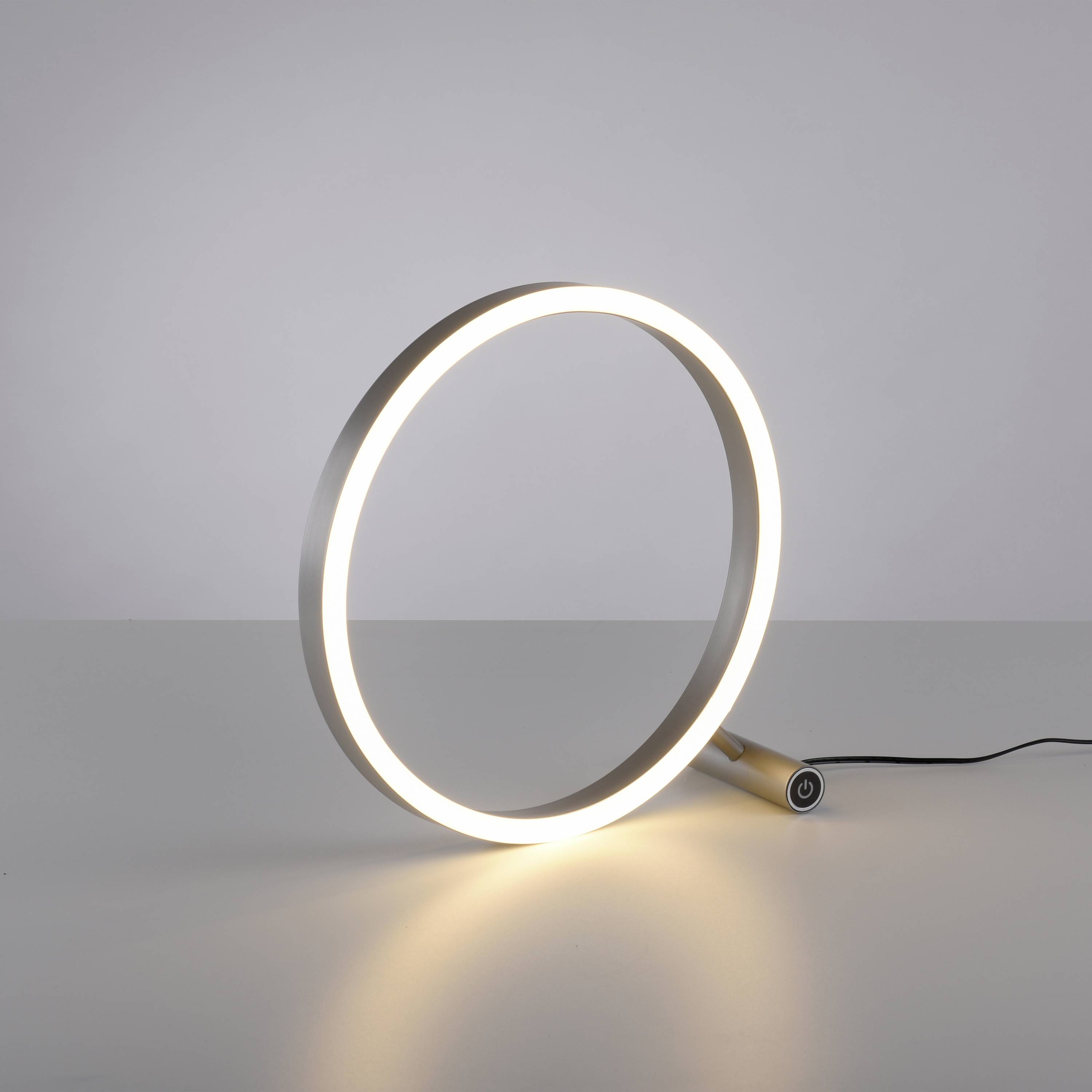 Eine moderne Tischlampe in Form eines leuchtenden Rings steht auf einem grauen Tisch. Sie leuchtet sanft und ist über ein Kabel verbunden.
