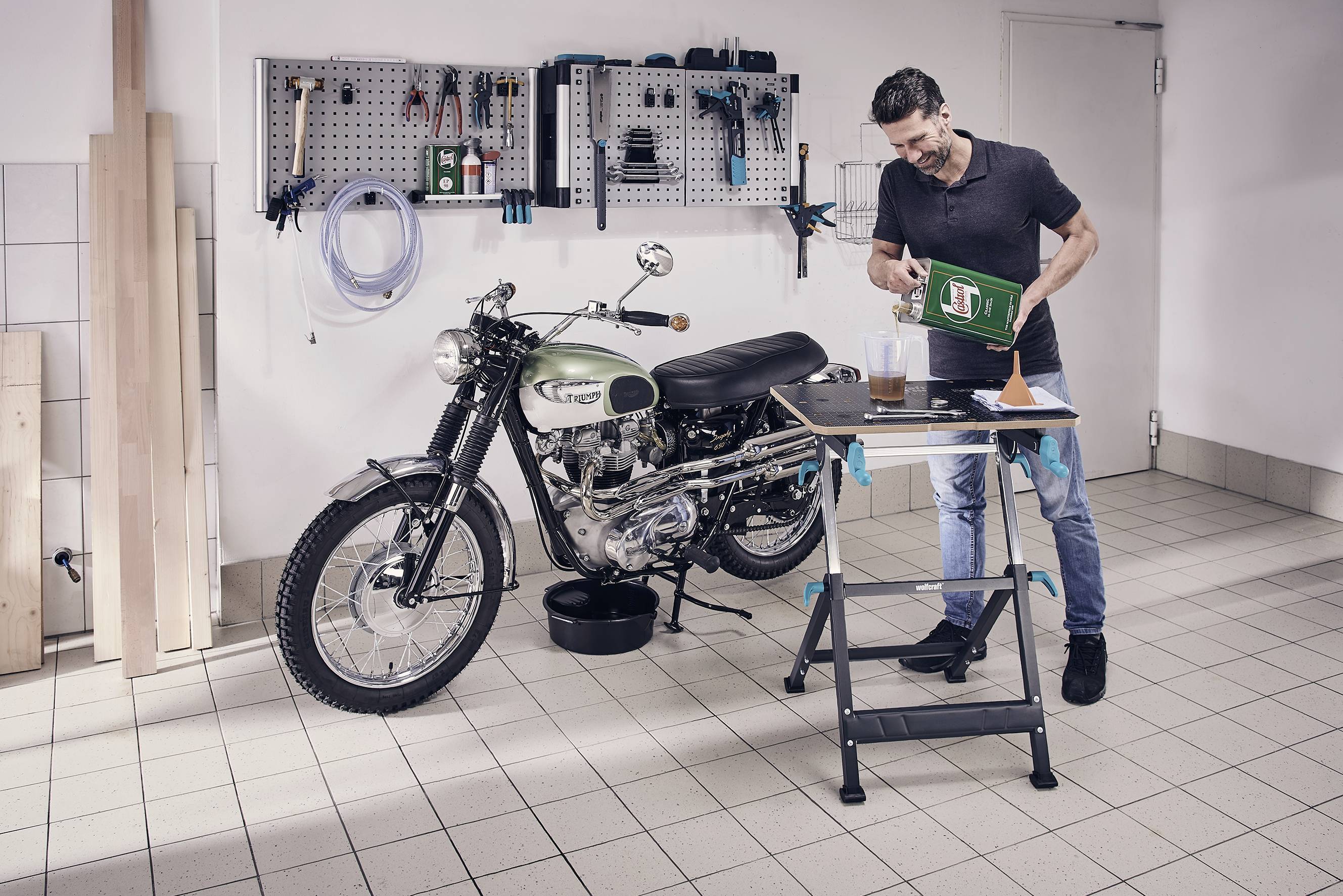 Mann in Werkstatt füllt Öl in ein Motorrad. Werkzeug und Zubehör an der Wand. Fokus auf Motorradwartung.