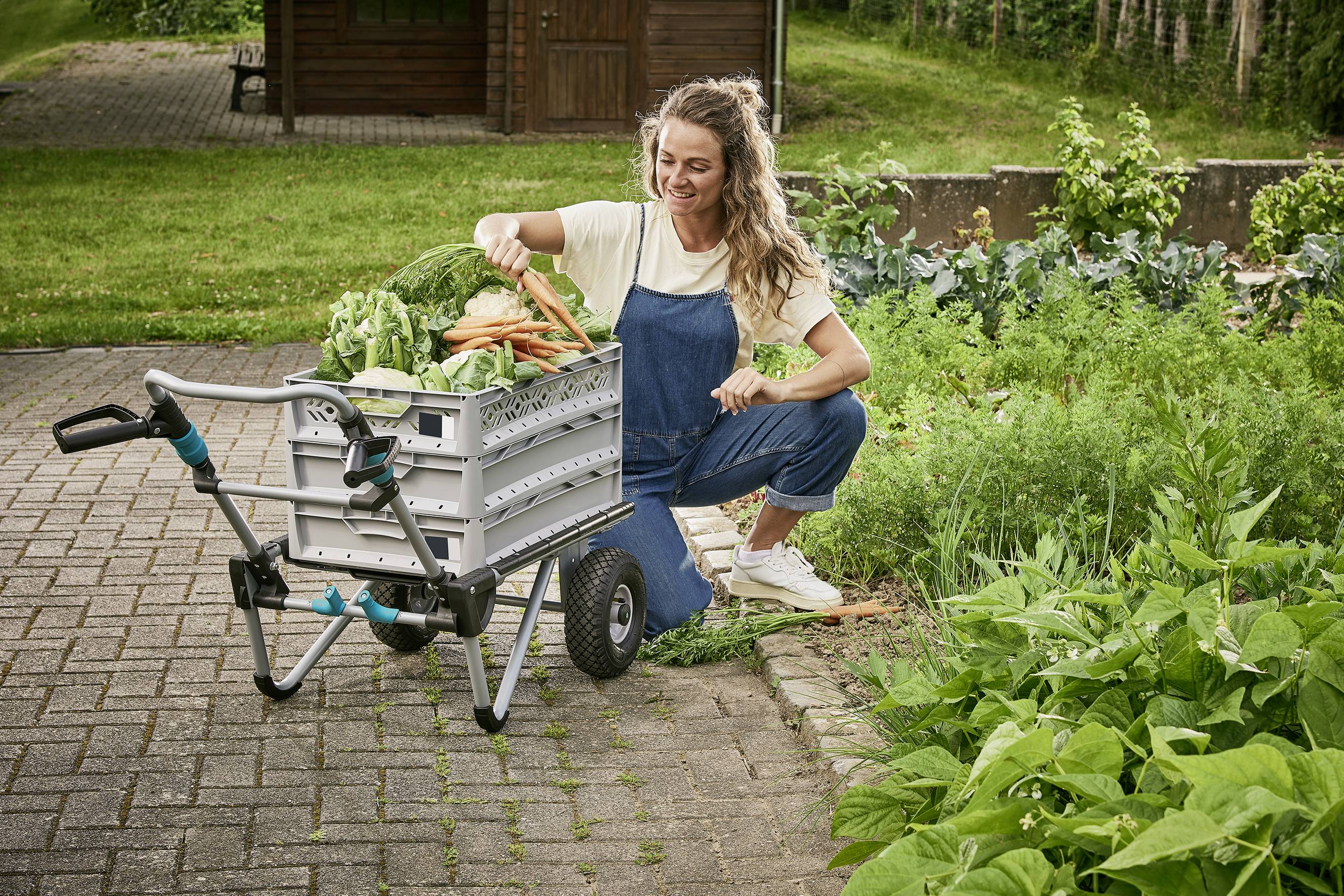 Frau in blauer Latzhose kniet und legt Gemüse, darunter Karotten und Kohl, vorsichtig in einen Gartenwagen. Im Hintergrund ein Garten.