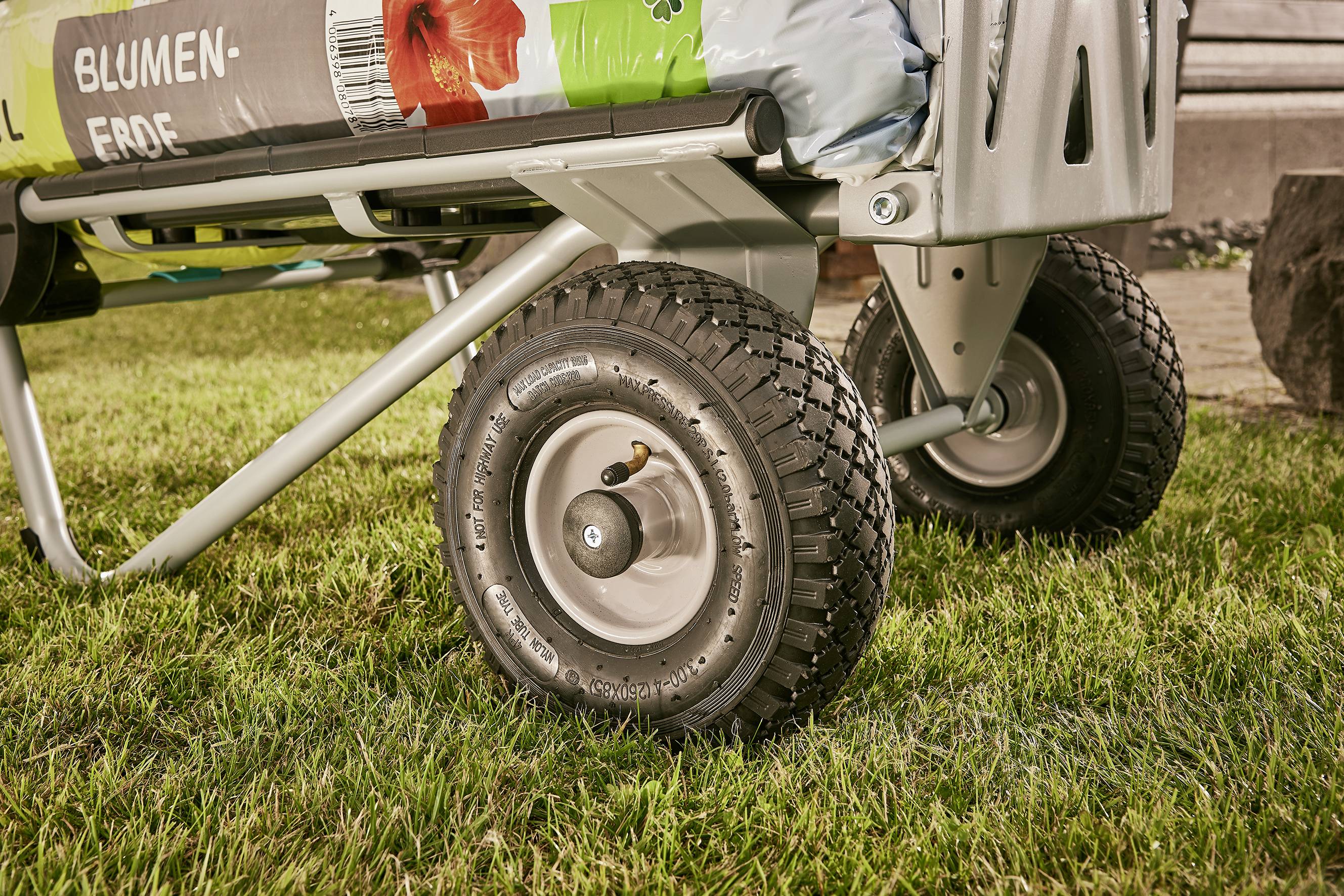 Nahaufnahme eines Metallkarren mit großen Reifen auf einer Wiese, beladen mit Säcken Blumenerde, die mit einer roten Blume bedruckt sind.