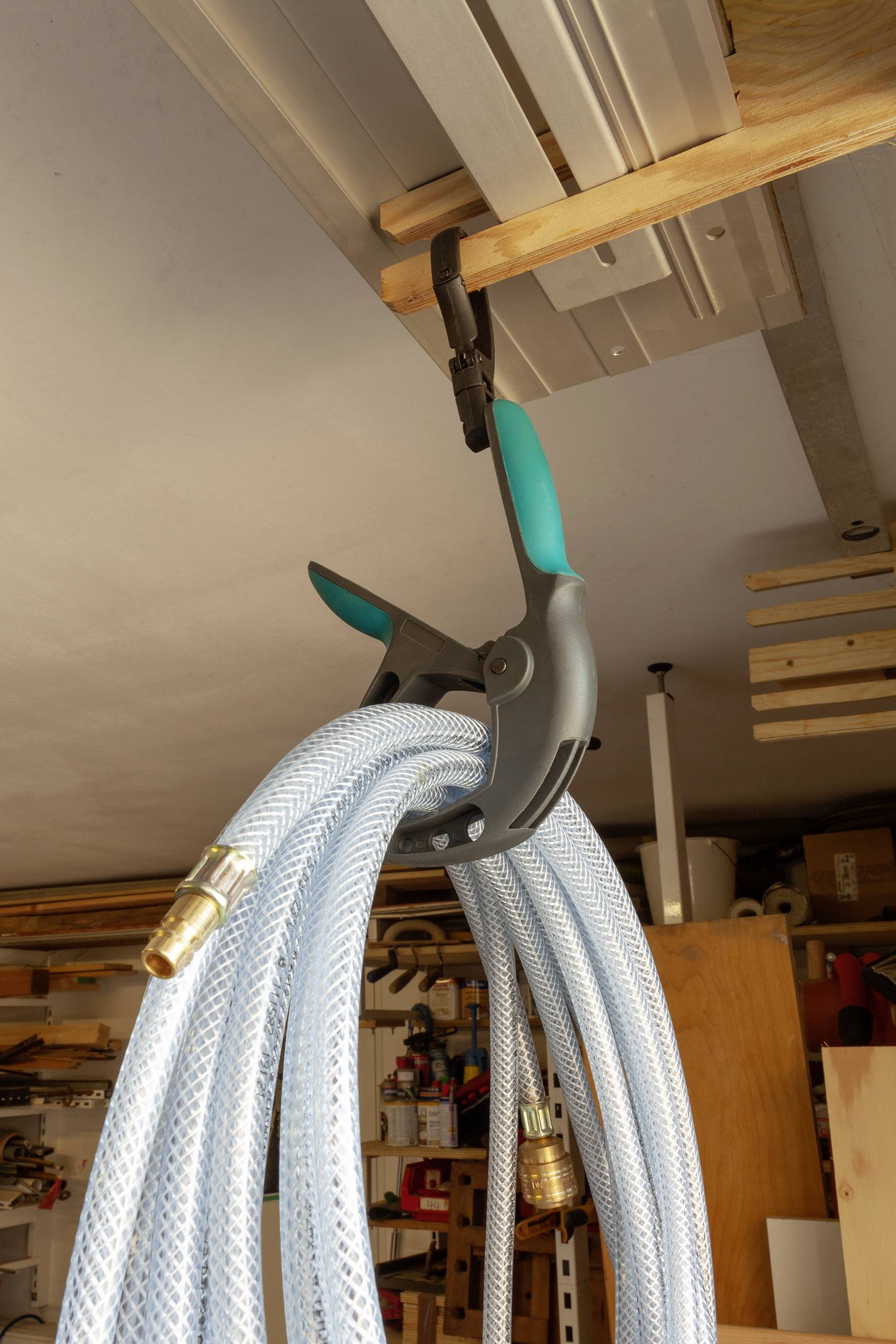 Ein grauer Schlauch wird mit einer großen Klemme an einem Holzregal in einer Werkstatt aufgehängt. Im Hintergrund sind Regale mit Werkzeugen zu sehen.