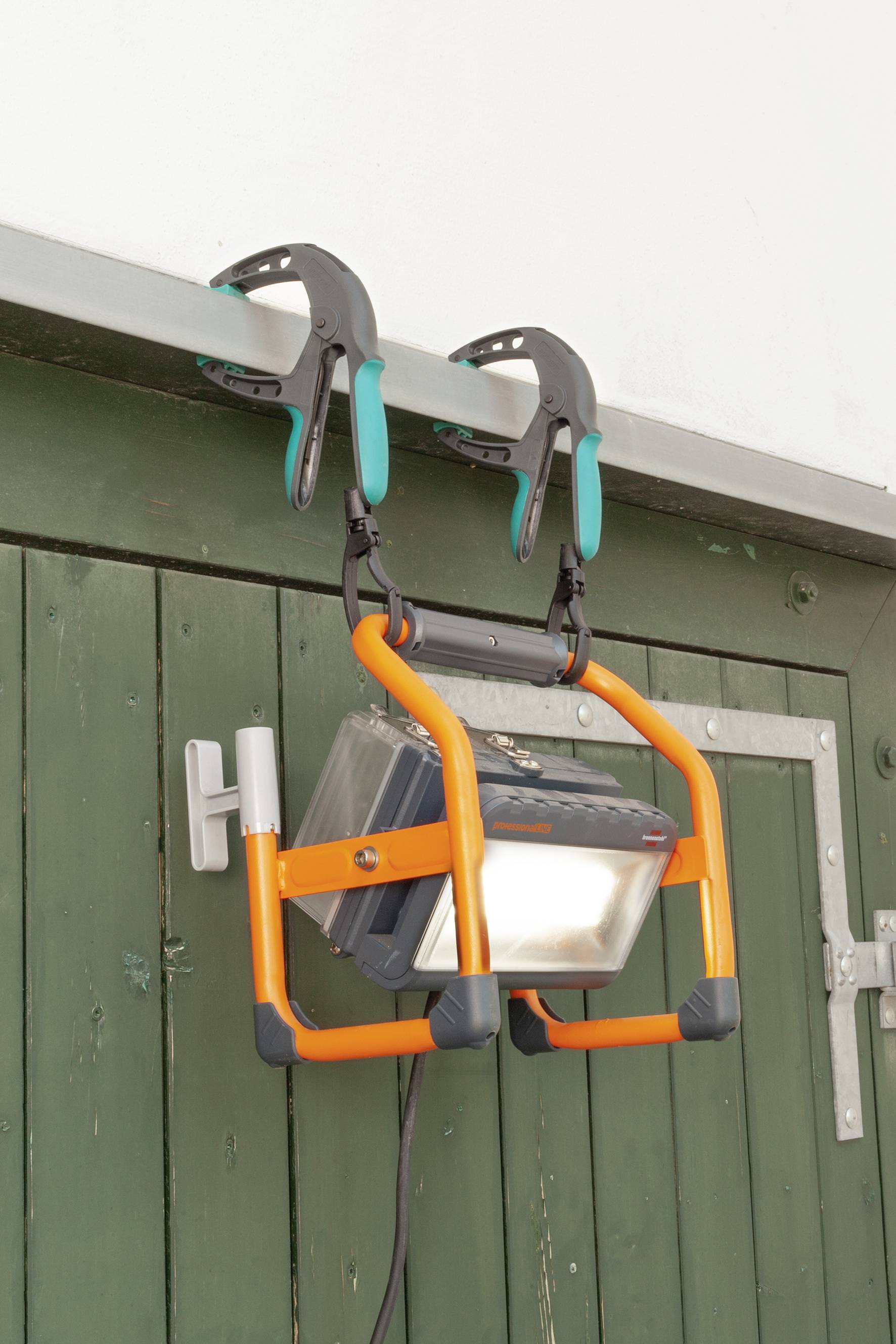 Eine tragbare Arbeitsleuchte ist mit zwei Klemmen an einer grünen Holztür befestigt. Sie hat einen orangefarbenen Rahmen.