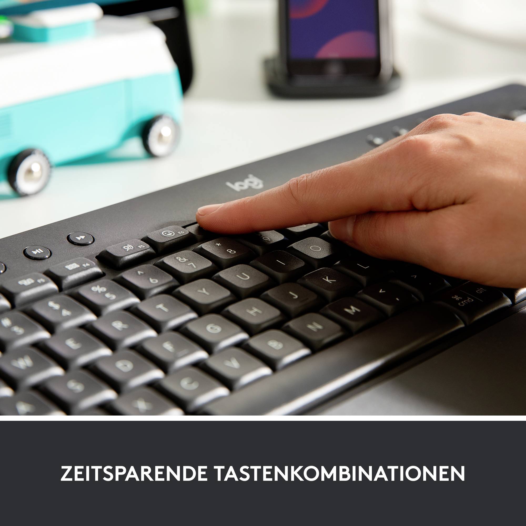 Eine Hand tippt auf einer Computertastatur. Im Hintergrund ist ein unscharfes Smartphone zu sehen.