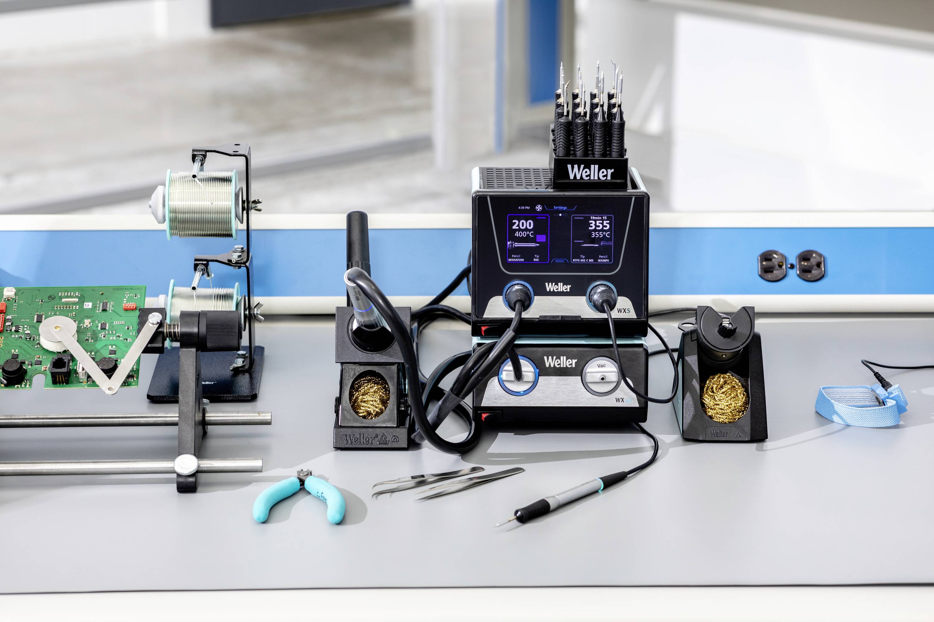 Elektronik-Arbeitsplatz mit Lötkolben, Leiterplatte in Halterung, Pinzette und Zangen. Hauptgerät zeigt Temperaturanzeige.