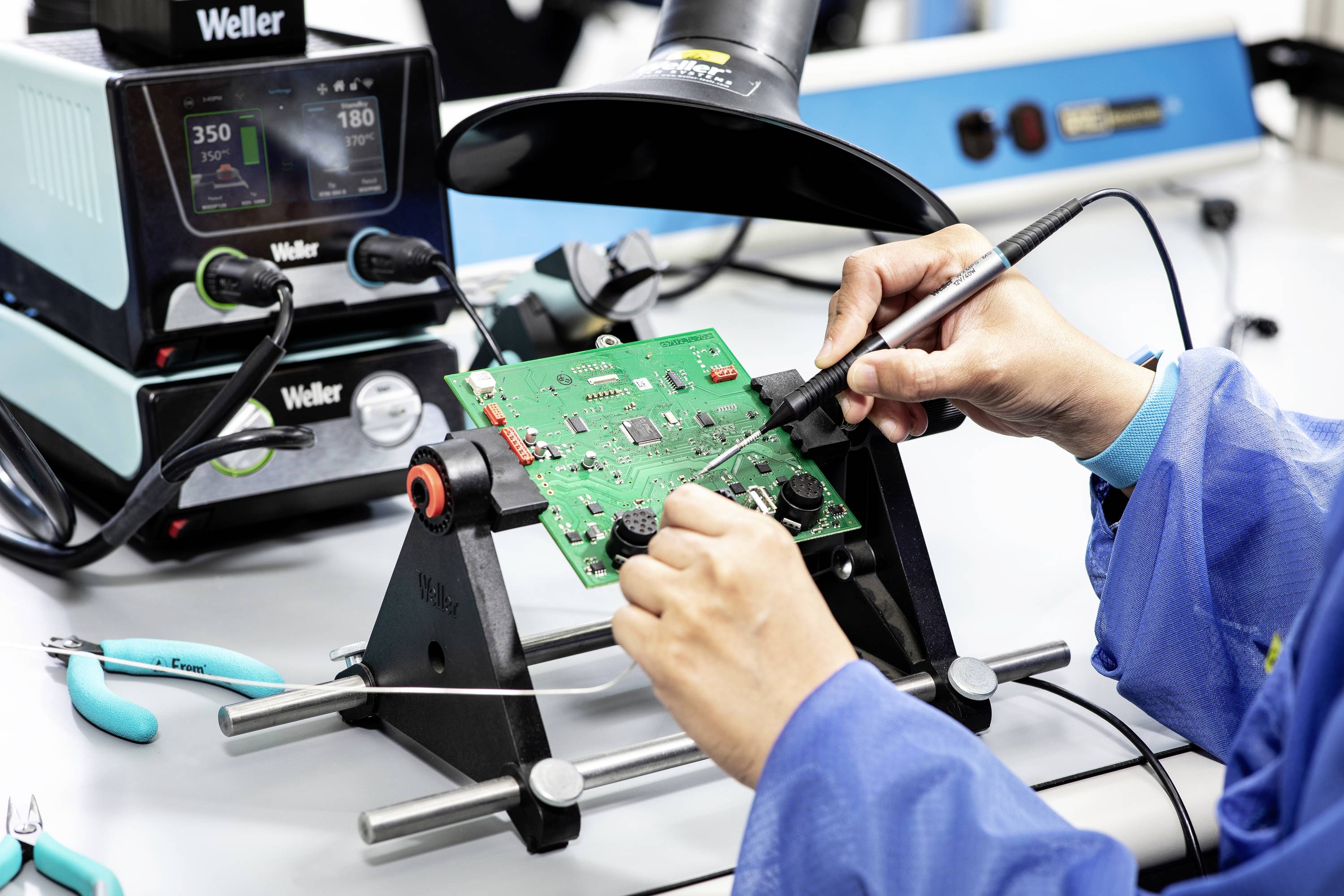 Person lötet an einer grünen Leiterplatte mit Werkzeugen. Im Hintergrund ein Lötkolben mit digitaler Anzeige.