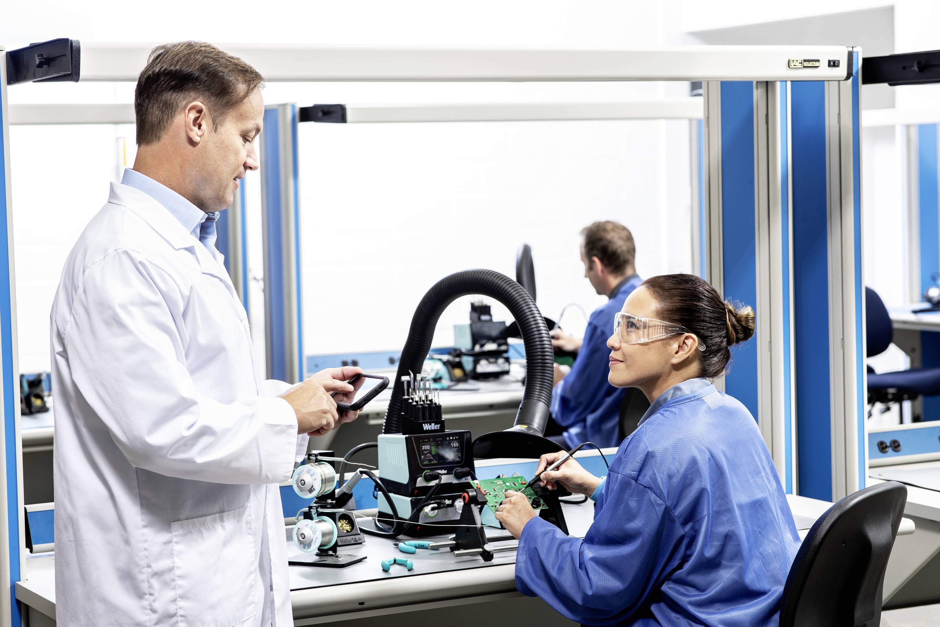 Ein Mann und eine Frau in Arbeitskitteln arbeiten an elektronischen Geräten in einem Labor. Sie kommunizieren miteinander.