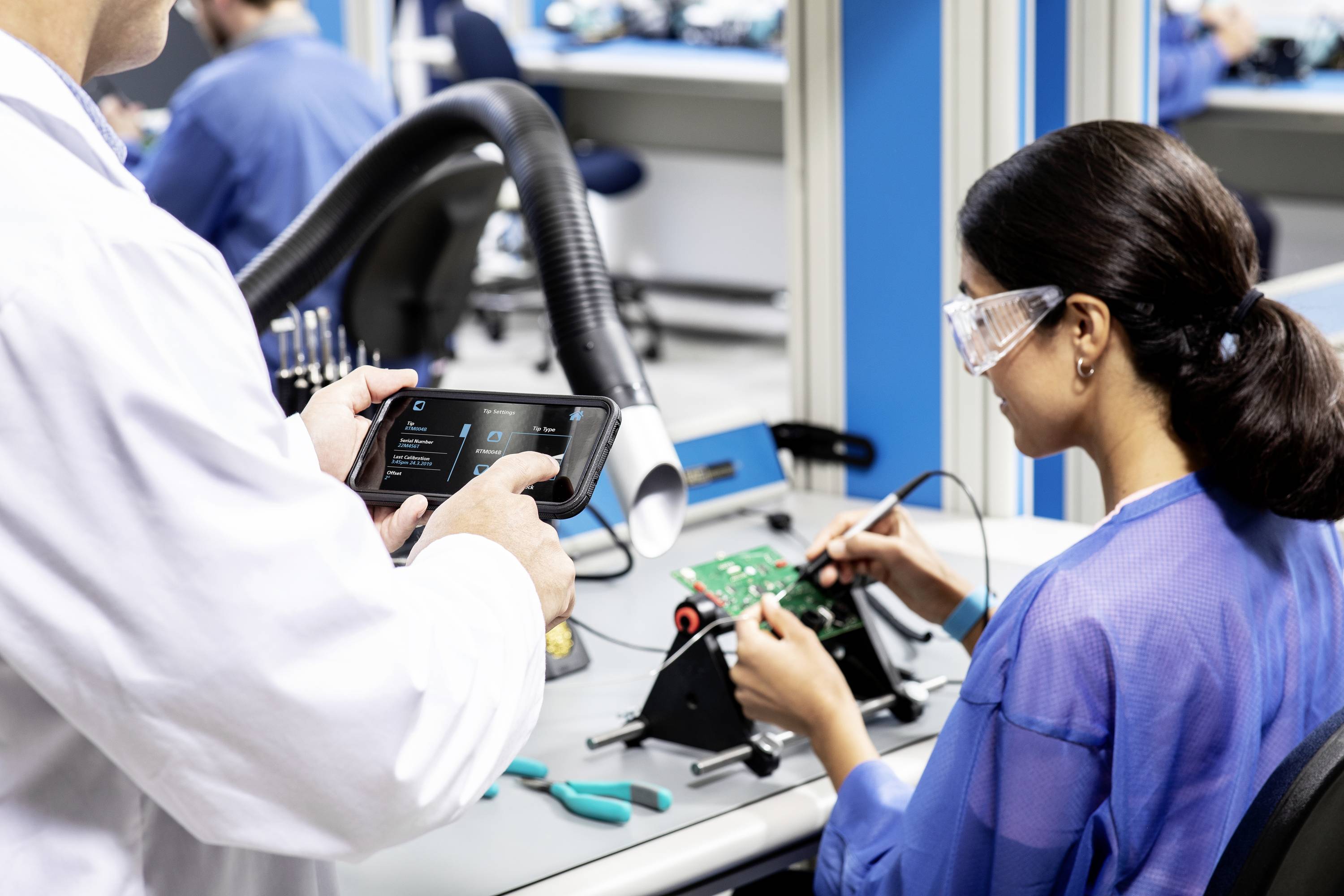 Zwei Personen in einem Labor. Eine Person arbeitet mit elektronischen Komponenten, während die andere ein Tablet für Messungen nutzt.