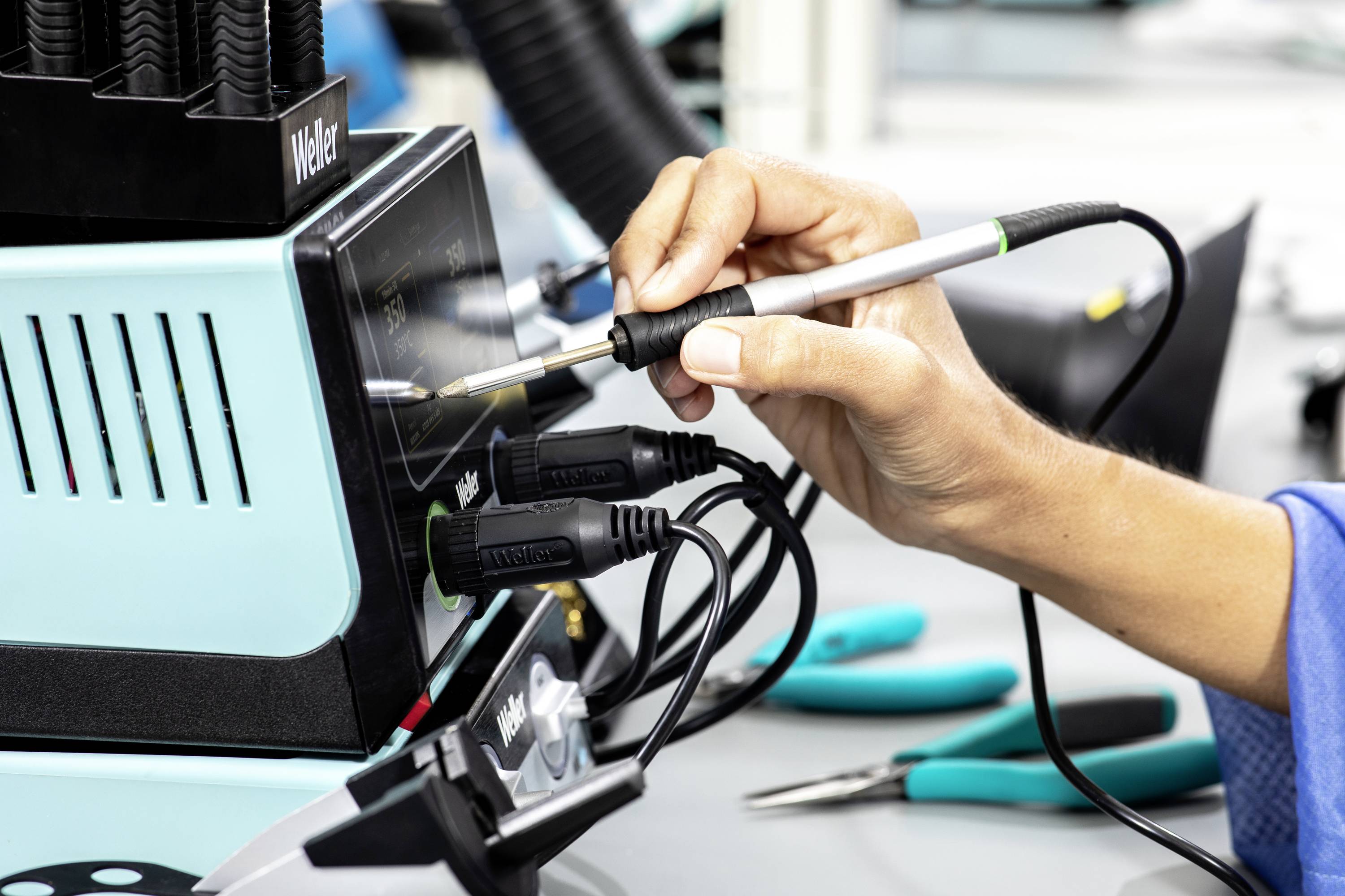 Eine Person verwendet einen Lötkolben an einer elektrischen Lötstation auf einem Arbeitstisch mit Werkzeugen.