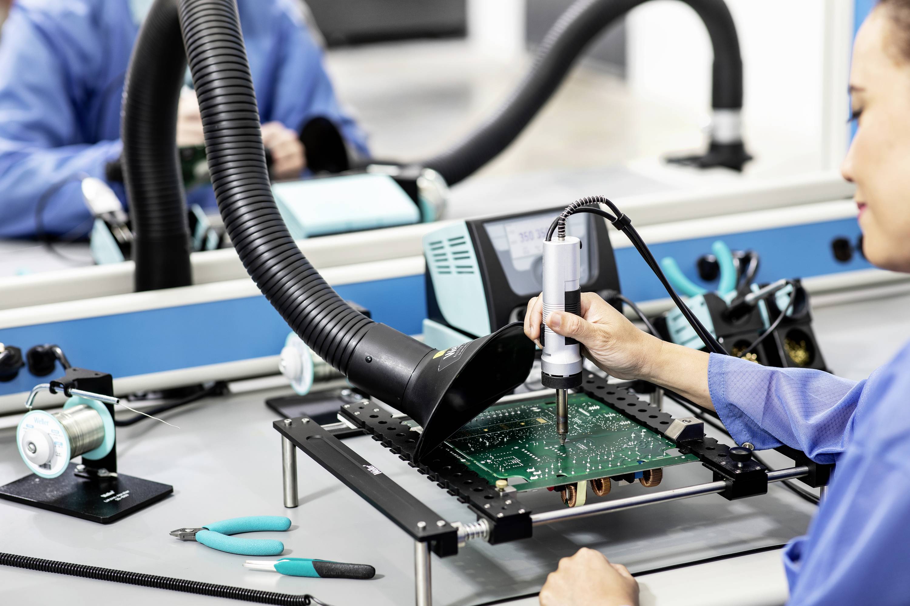 Ingenieurin lötet eine Platine in einer Elektronikwerkstatt, umgeben von technischen Geräten und Werkzeugen wie Zangen und Lötkolben.