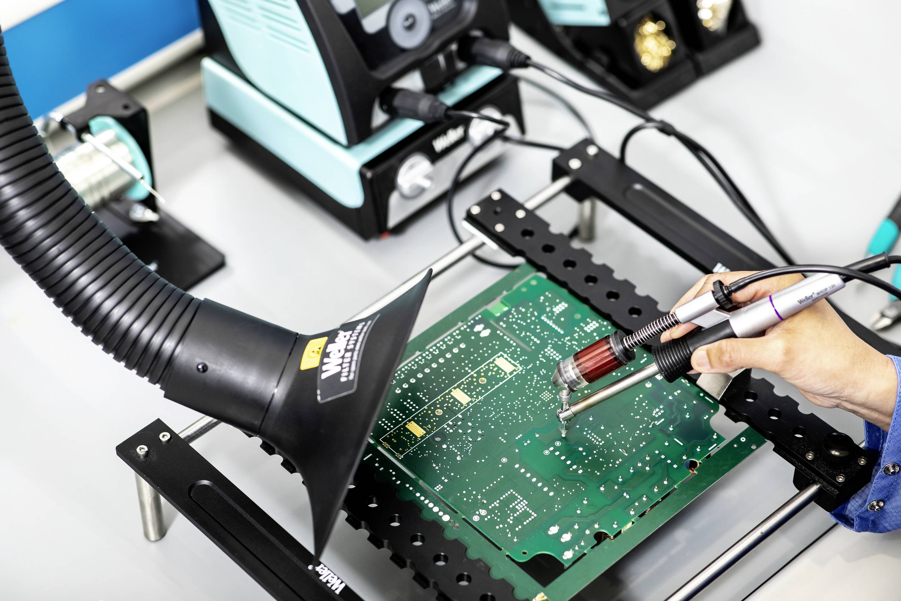 Eine Person lötet eine Leiterplatte, wobei ein Absaugarm in der Nähe ist. Im Hintergrund ist Lötausrüstung zu sehen.