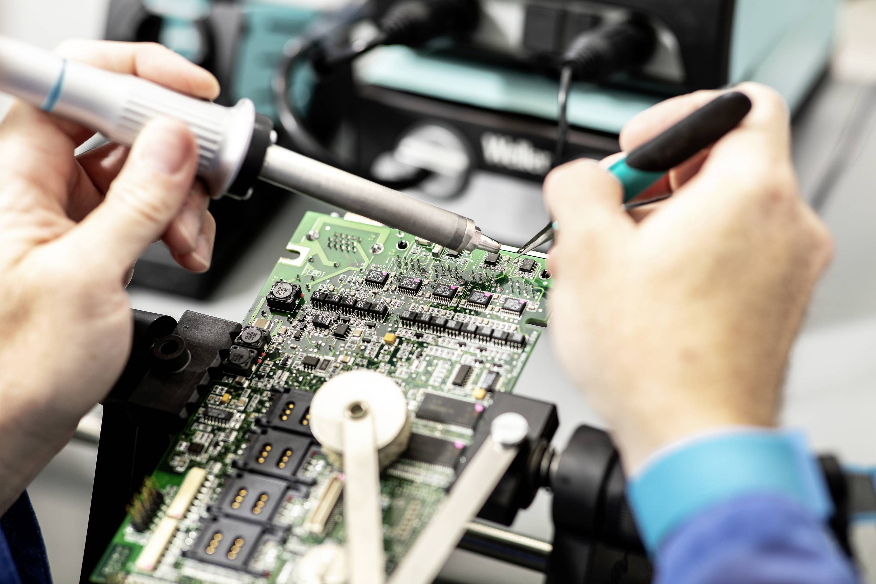 Eine Person lötet präzise an einer elektronischen Leiterplatte, umgeben von technischen Geräten, in einem Laborumfeld.