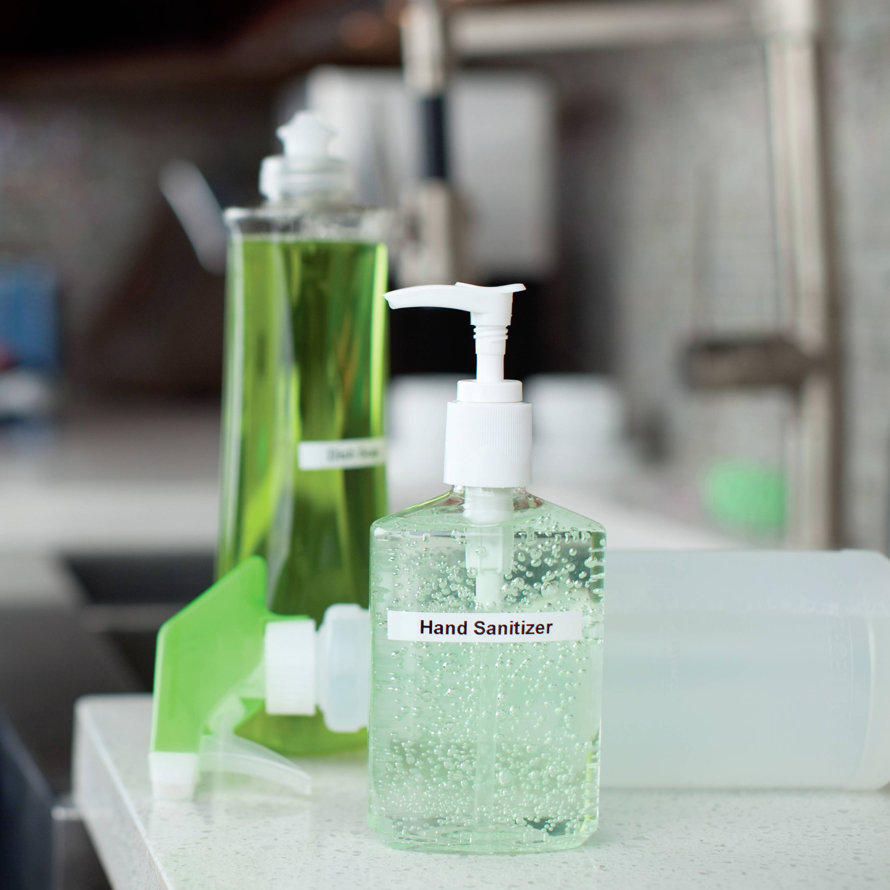 Im Vordergrund steht ein durchsichtiger Spender mit 'Hand Sanitizer', gefüllt mit einer klaren Flüssigkeit, in einer Küche.
