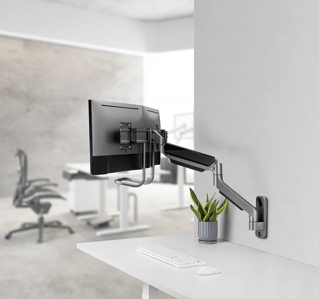 Ein Computermonitor ist an einem verstellbaren Wandarm befestigt. Darunter befinden sich eine Tastatur und eine kleine Pflanze.
