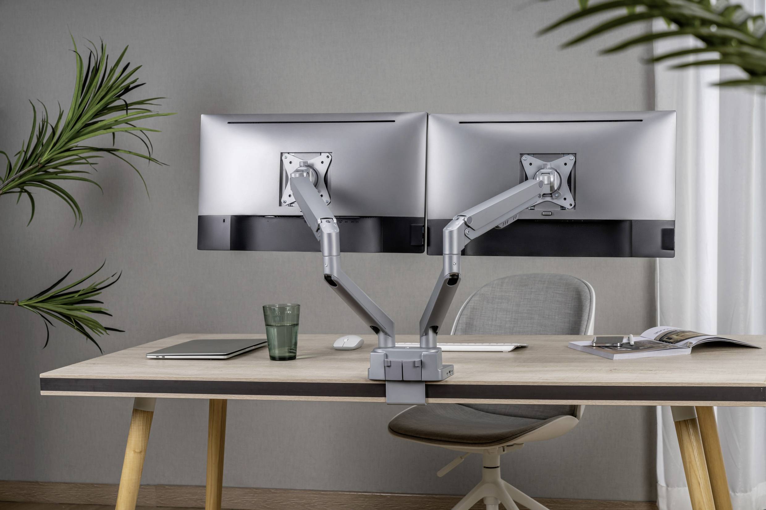 Ein moderner Schreibtisch mit zwei großen Monitoren auf verstellbaren Halterungen, einem Laptop, Glas, und einer Zeitschrift.