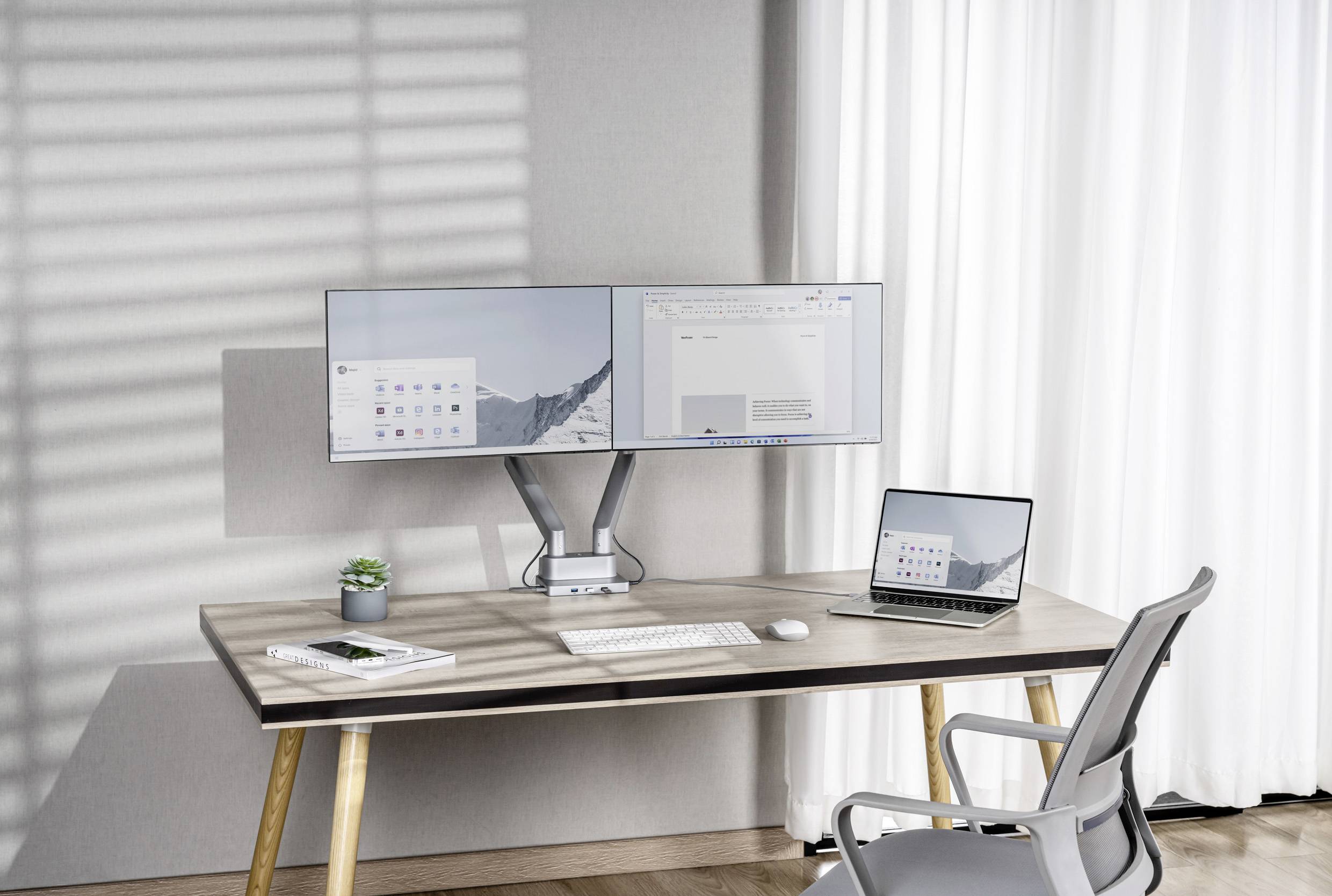 Ein Schreibtisch mit zwei Monitoren, einem Laptop, Tastatur und Maus. Im Hintergrund große Fenster mit weißen Vorhängen.