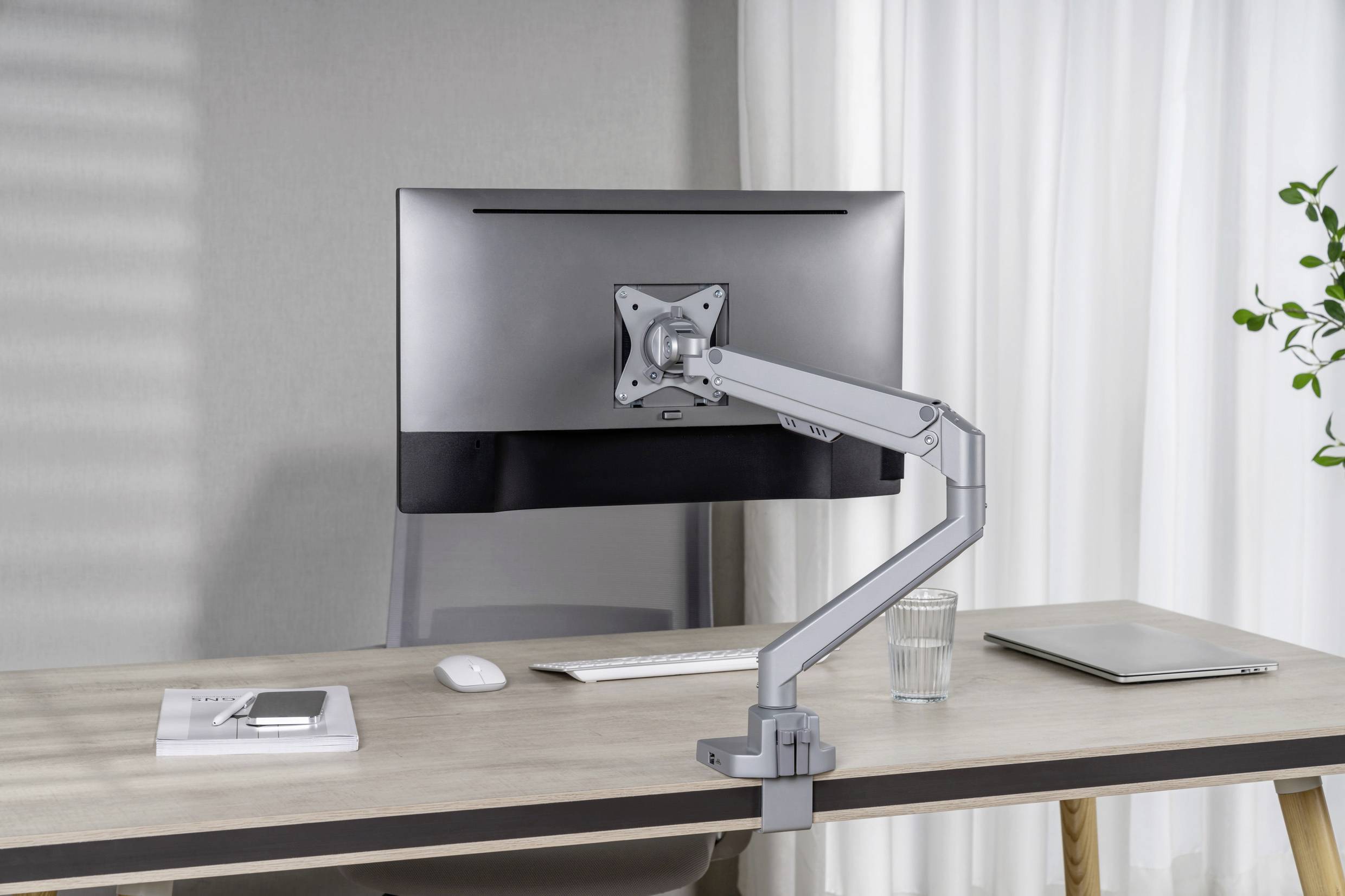 Ein Schreibtisch mit einem Monitor auf einem verstellbaren Arm, eine Tastatur, ein Glas Wasser, ein Laptop und ein Buch mit Smartphone liegen darauf.