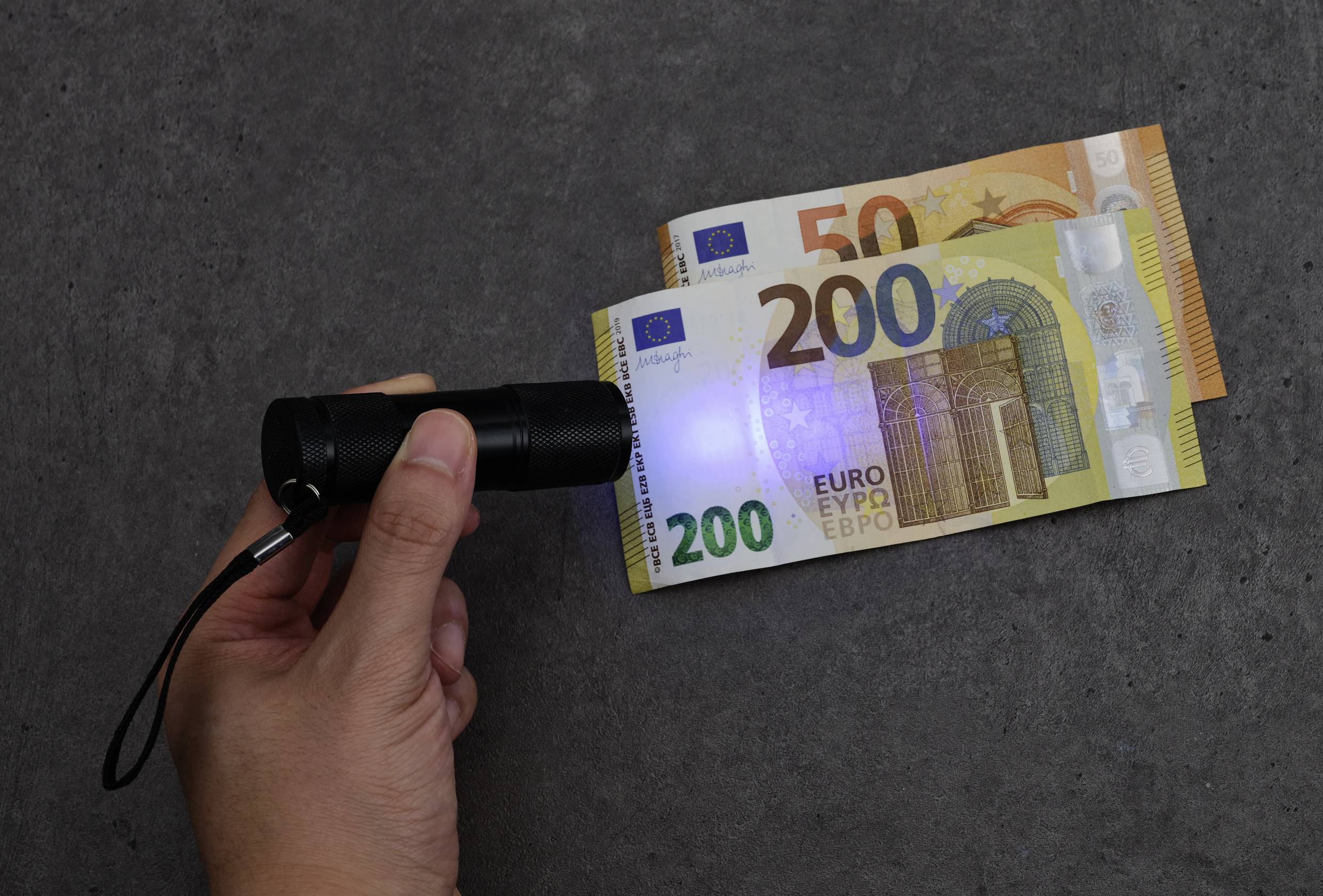 Eine Hand hält eine UV-Taschenlampe über zwei Euro-Geldscheine (200 und 50 Euro), um Sicherheitsmerkmale sichtbar zu machen.