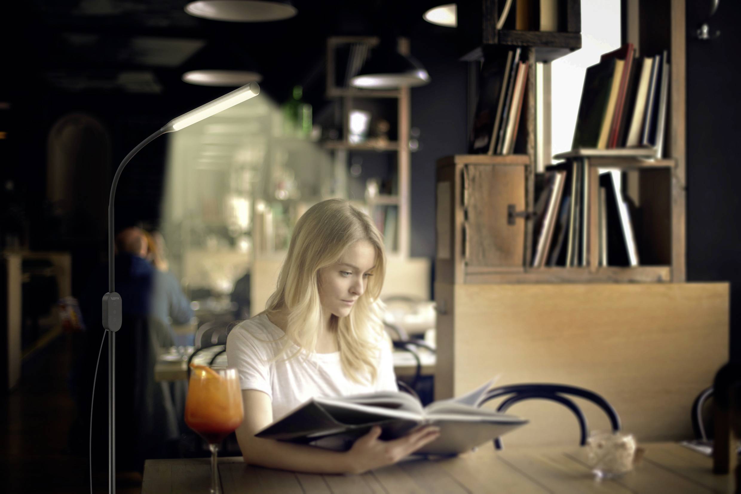 Eine Frau liest in einem gemütlichen Café. Auf dem Tisch steht ein Getränk. Im Hintergrund sind Bücherregale sichtbar.