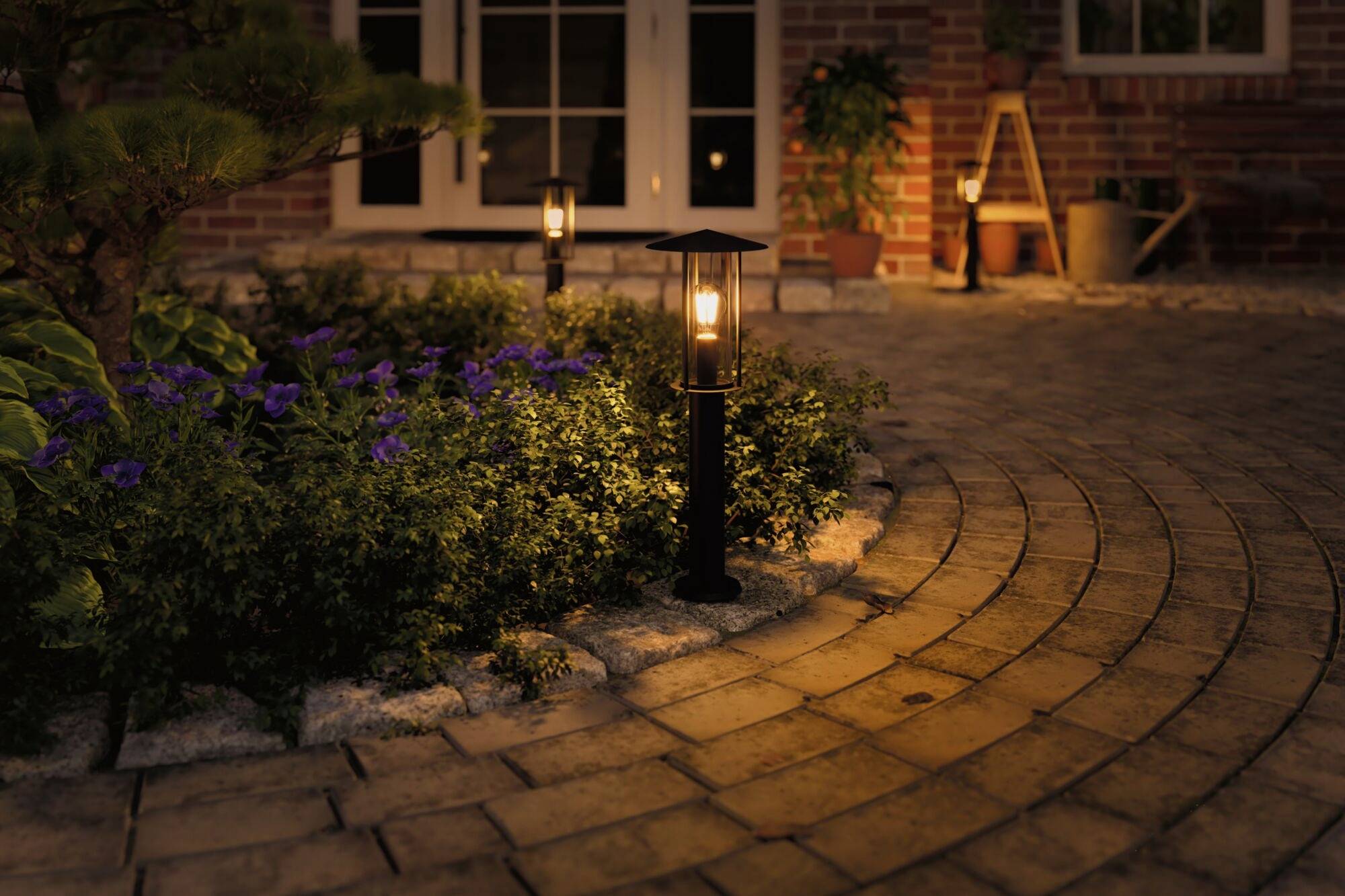 Gepflasterter Gartenweg bei Nacht, beleuchtet von Laternen, umgeben von blühenden Pflanzen vor einem Haus mit Ziegelwänden.
