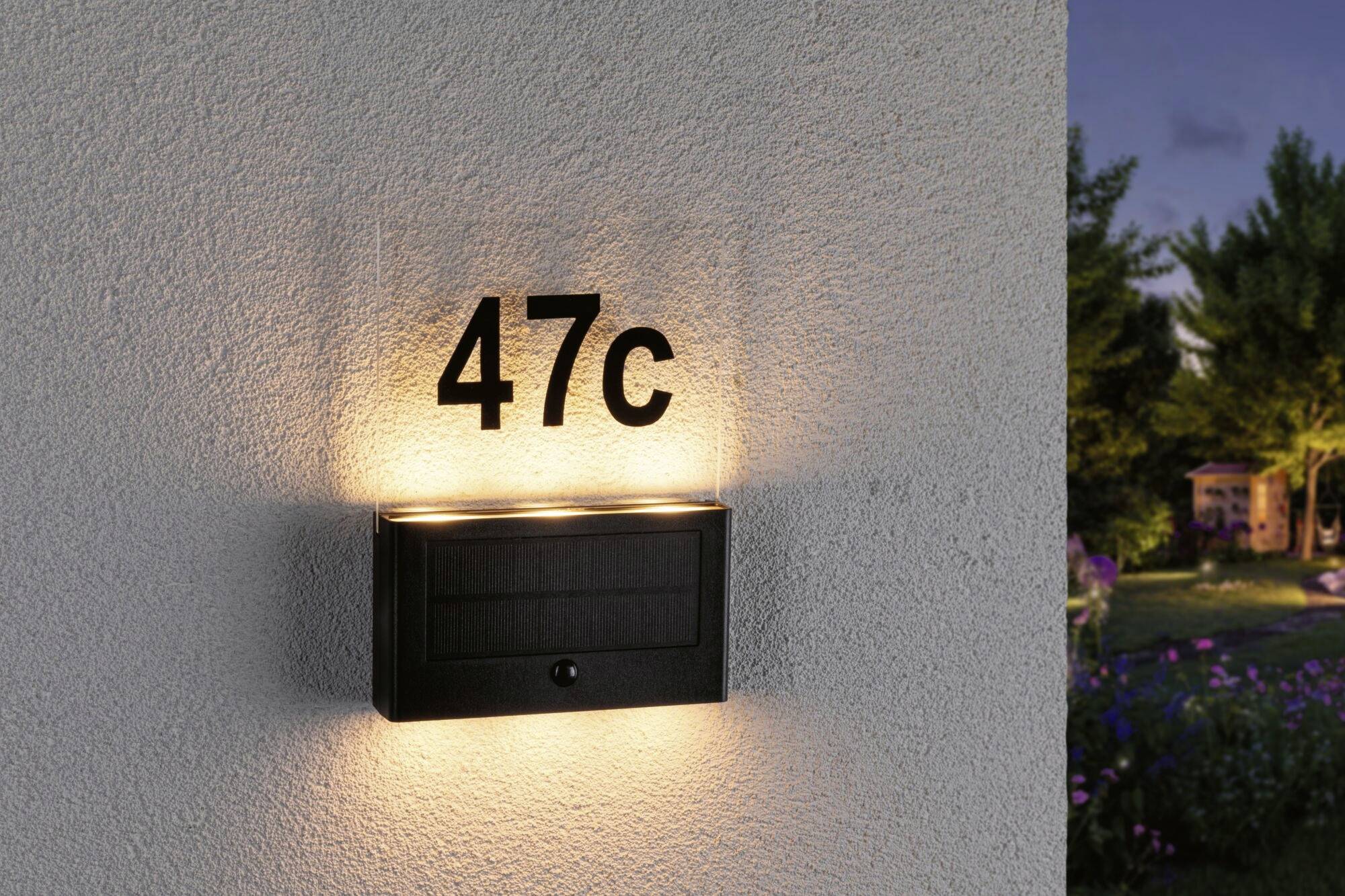 Beleuchtete Hausnummer '47c' auf einer Wand bei Dämmerung, umgeben von Garten und Bäumen.