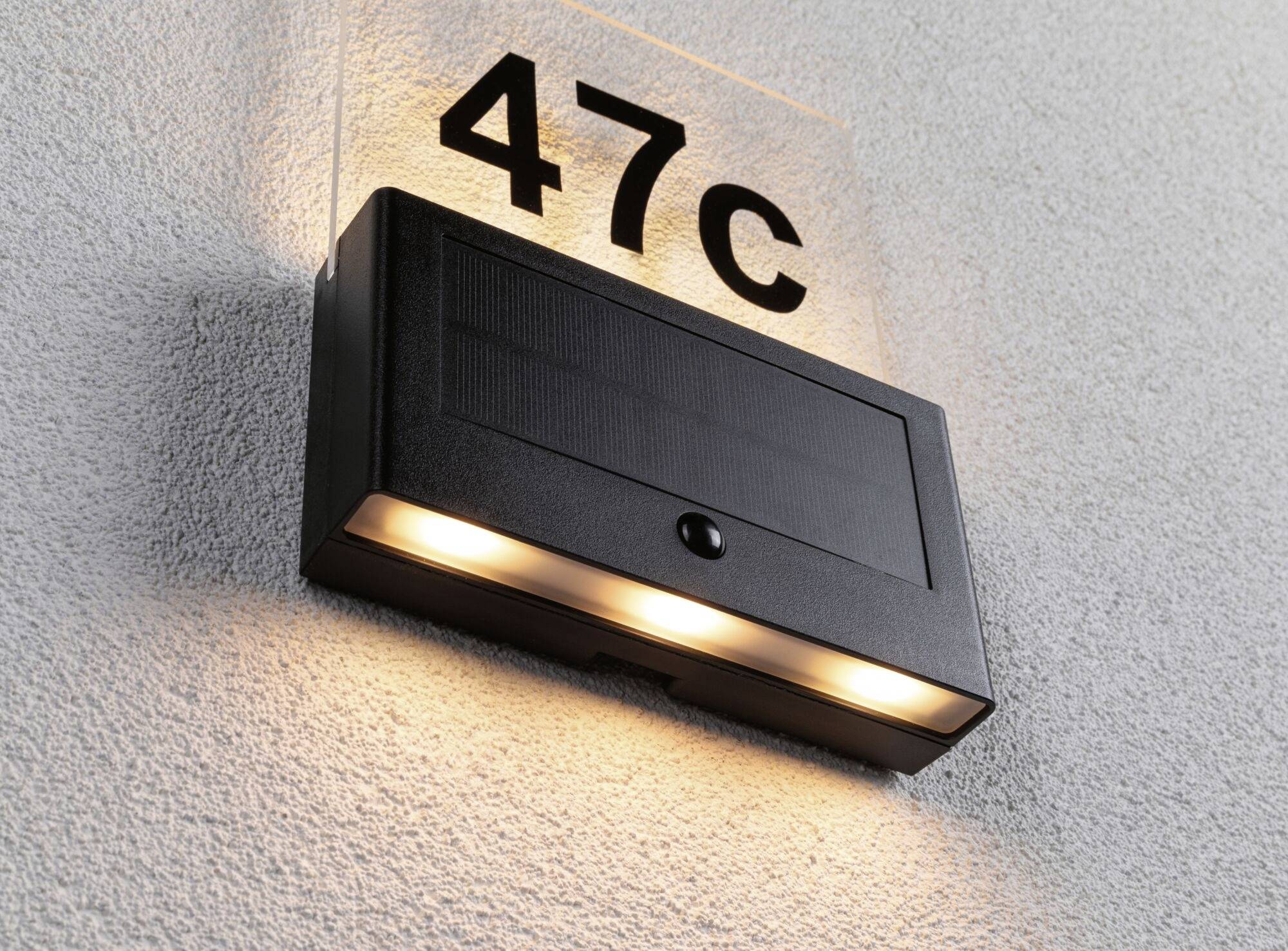 An einer grauen Wand hängt eine schwarze Außenlampe mit drei leuchtenden Lampen unterhalb eines Schilds mit der Aufschrift '47C'.