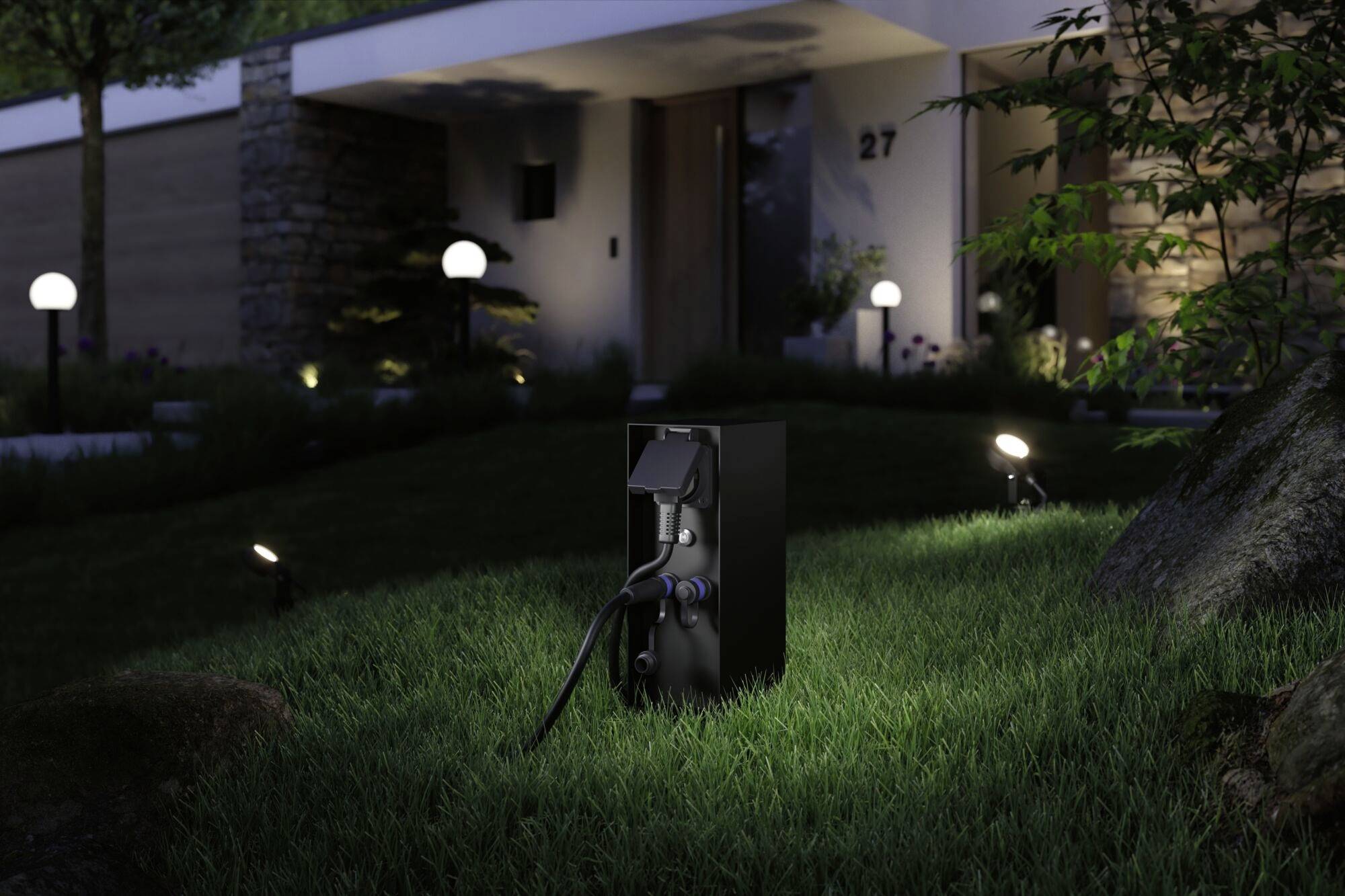 Eine schwarze Ladestation für Elektroautos steht auf einem Rasen vor einem modernen Haus bei Nacht, beleuchtet von kleinen Bodenlichtern.