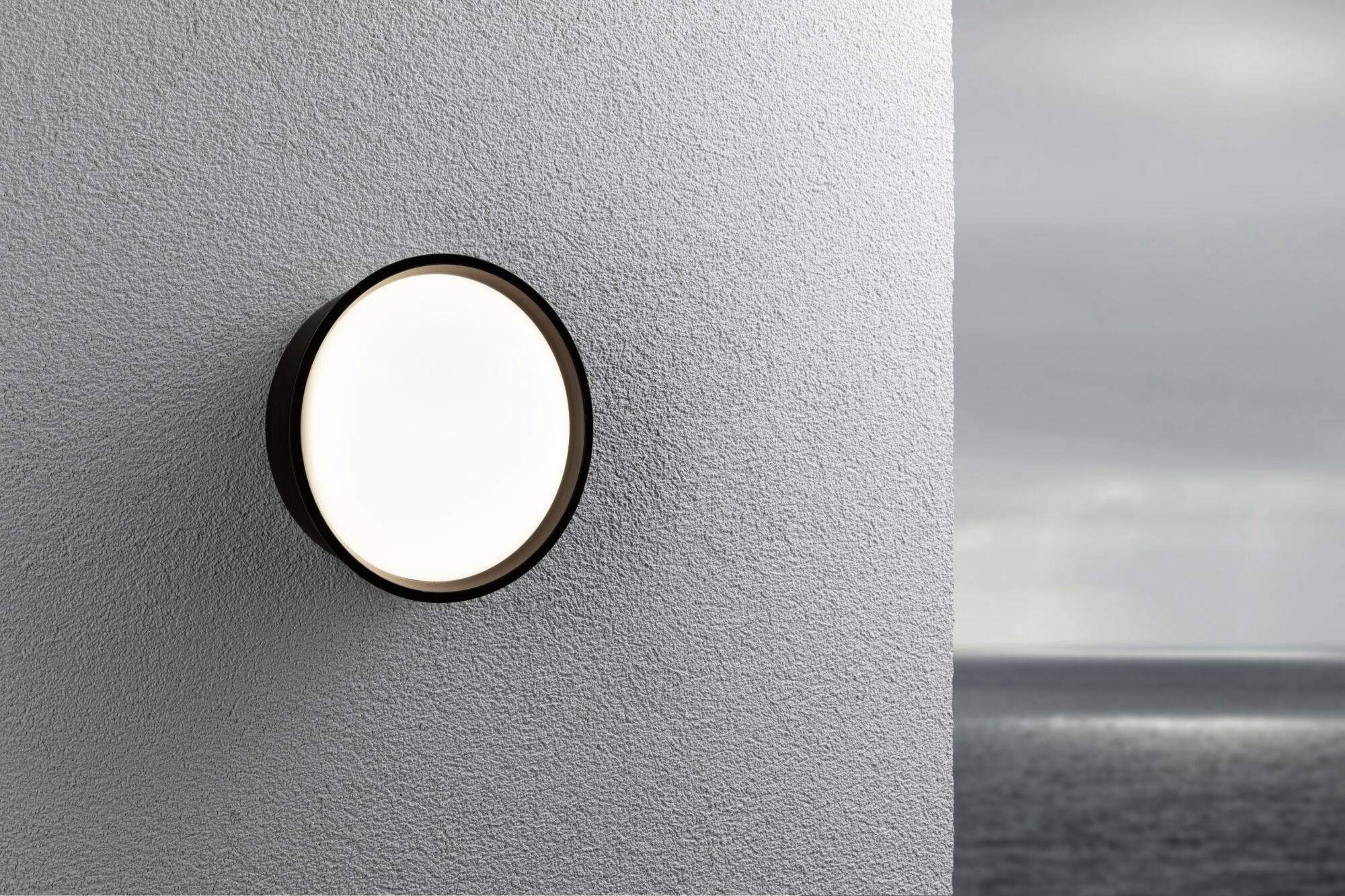 Eine runde, moderne Außenleuchte an einer grauen Wand leuchtet. Im Hintergrund ist ein bewölkter Himmel über dem Meer sichtbar.