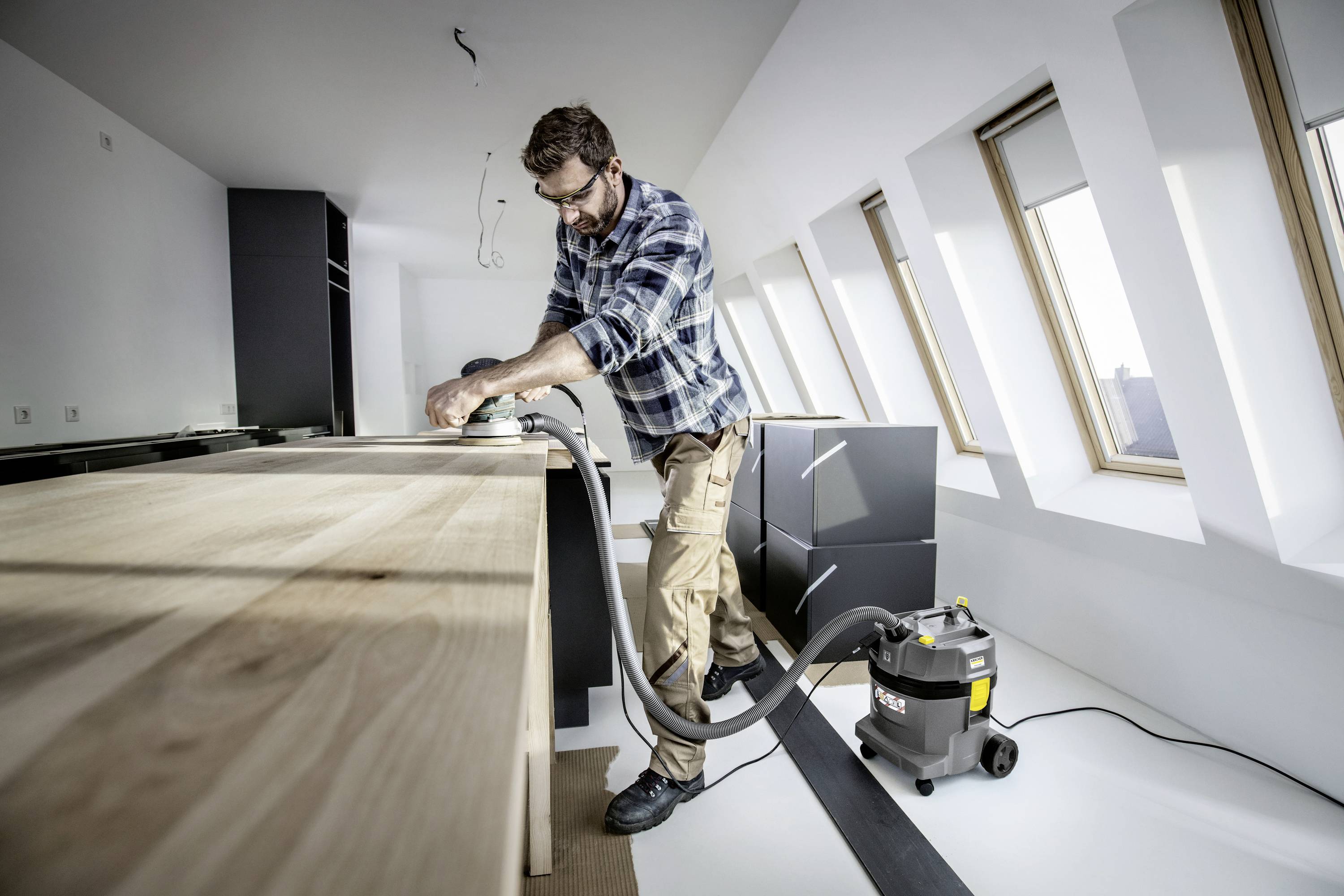 Ein Mann schleift eine Holzplatte in einer modernen, hellen Küche. Ein Staubsauger steht neben ihm, um den Staub aufzufangen.